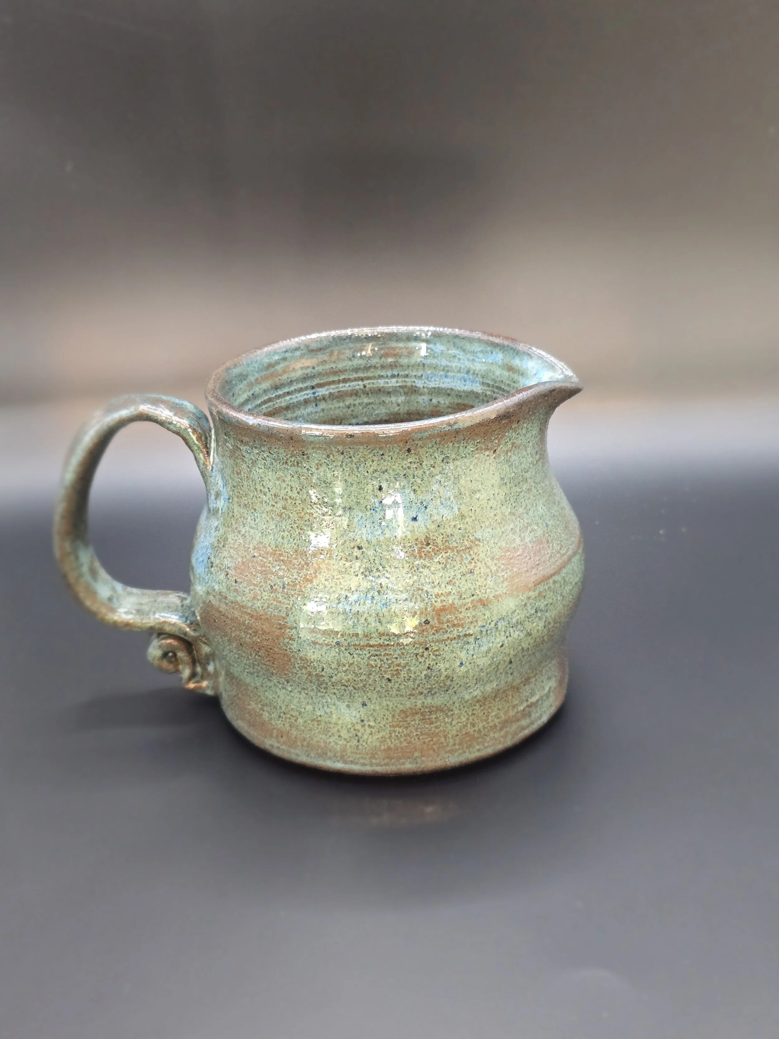 Ceramic mug with a glaze finish, featuring earthy tones of green, blue, and brown, placed on a black surface with a blurred gray background.