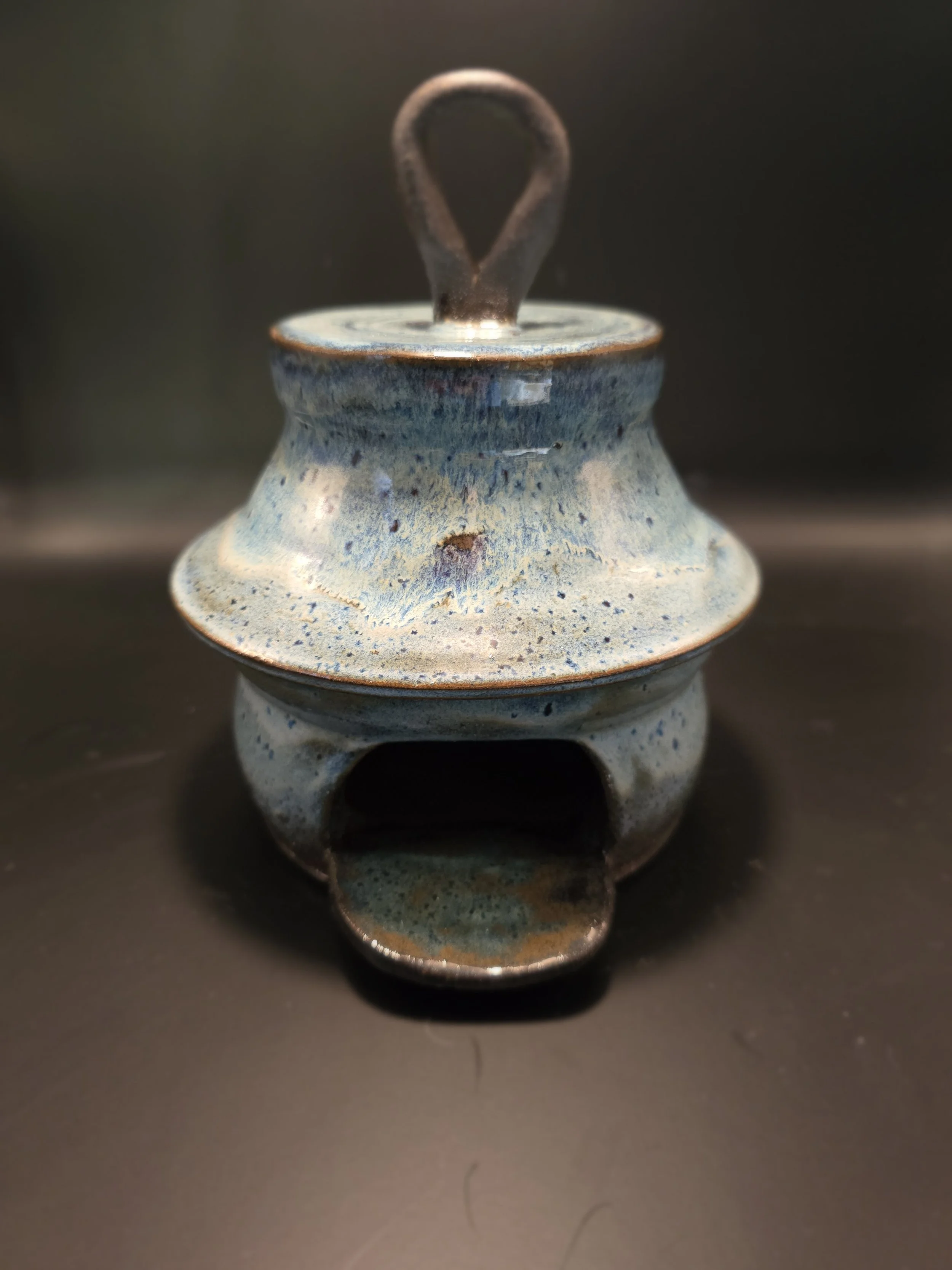 A ceramic incense burner with a lid and a handle, glazed in blue and earth tones, sitting on a dark surface.