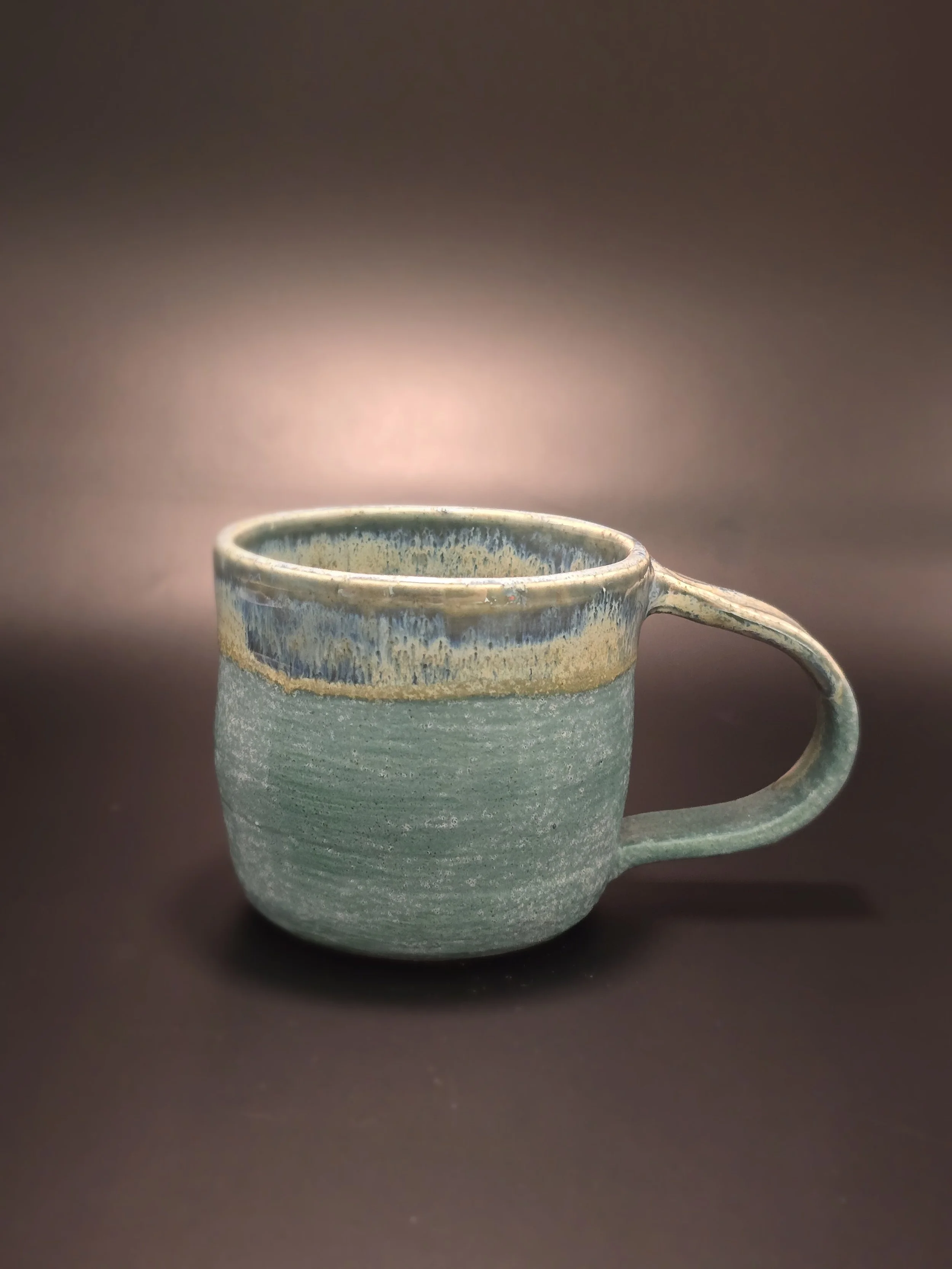 Close-up of a handmade ceramic mug with a textured green body and a blue and beige glazed rim, set against a dark blurred background.
