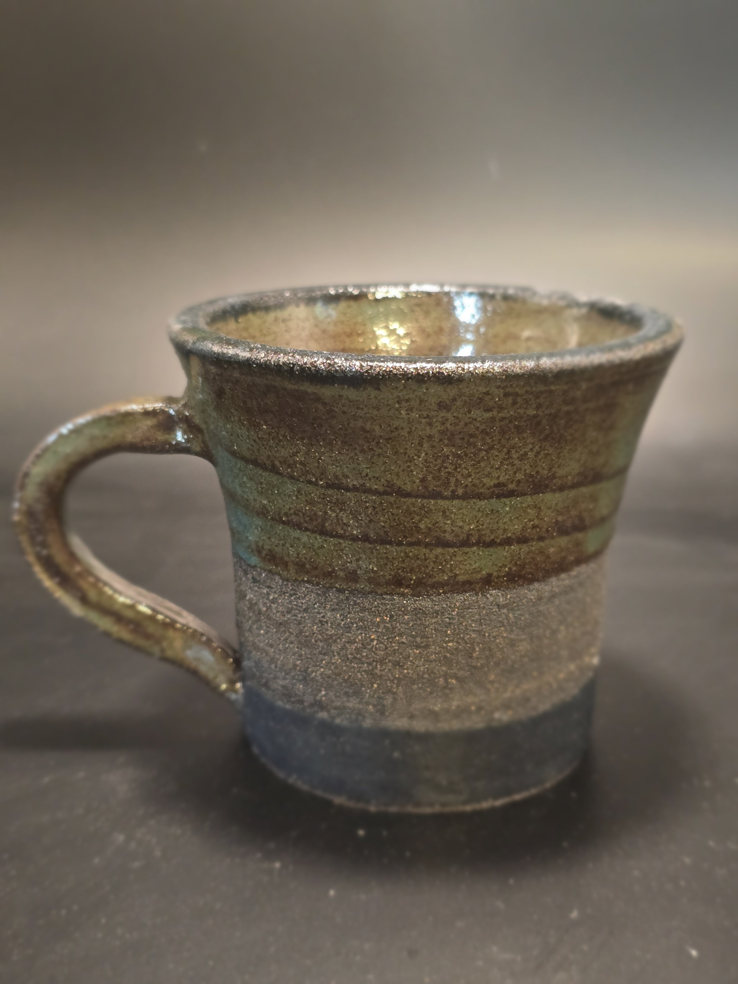 A ceramic mug with a textured glaze featuring horizontal lines and a blend of earthy tones including green, brown, and gray, placed on a dark surface with a blurred background.
