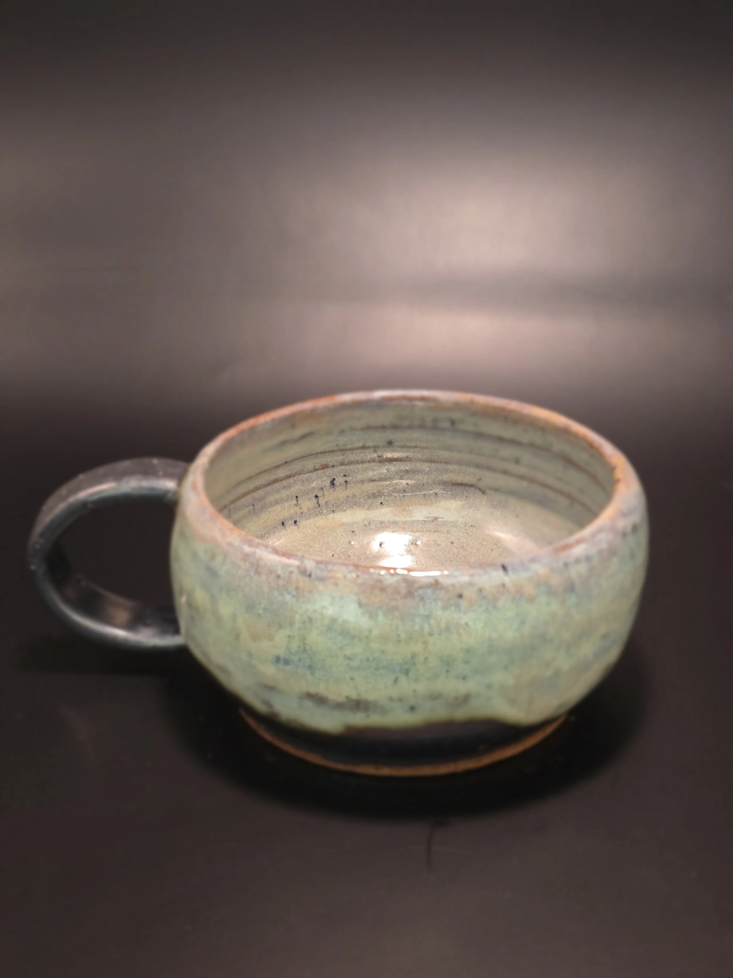 A ceramic cup with a round shape, handle on the side, glazed in earthy green and brown tones, placed on a dark surface.