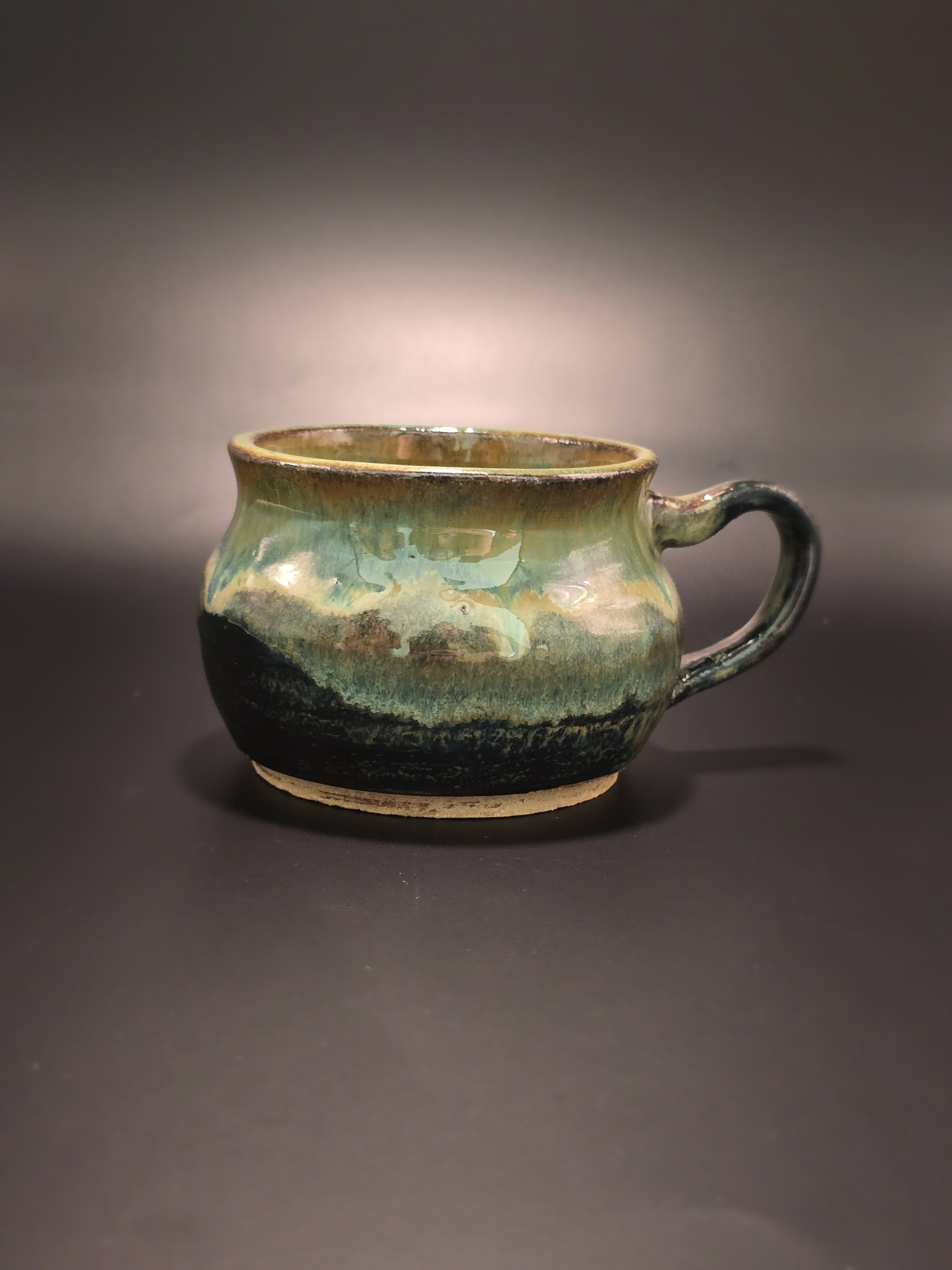 A ceramic mug with a rounded shape, green and black glaze, and a small handle, placed on a dark surface.