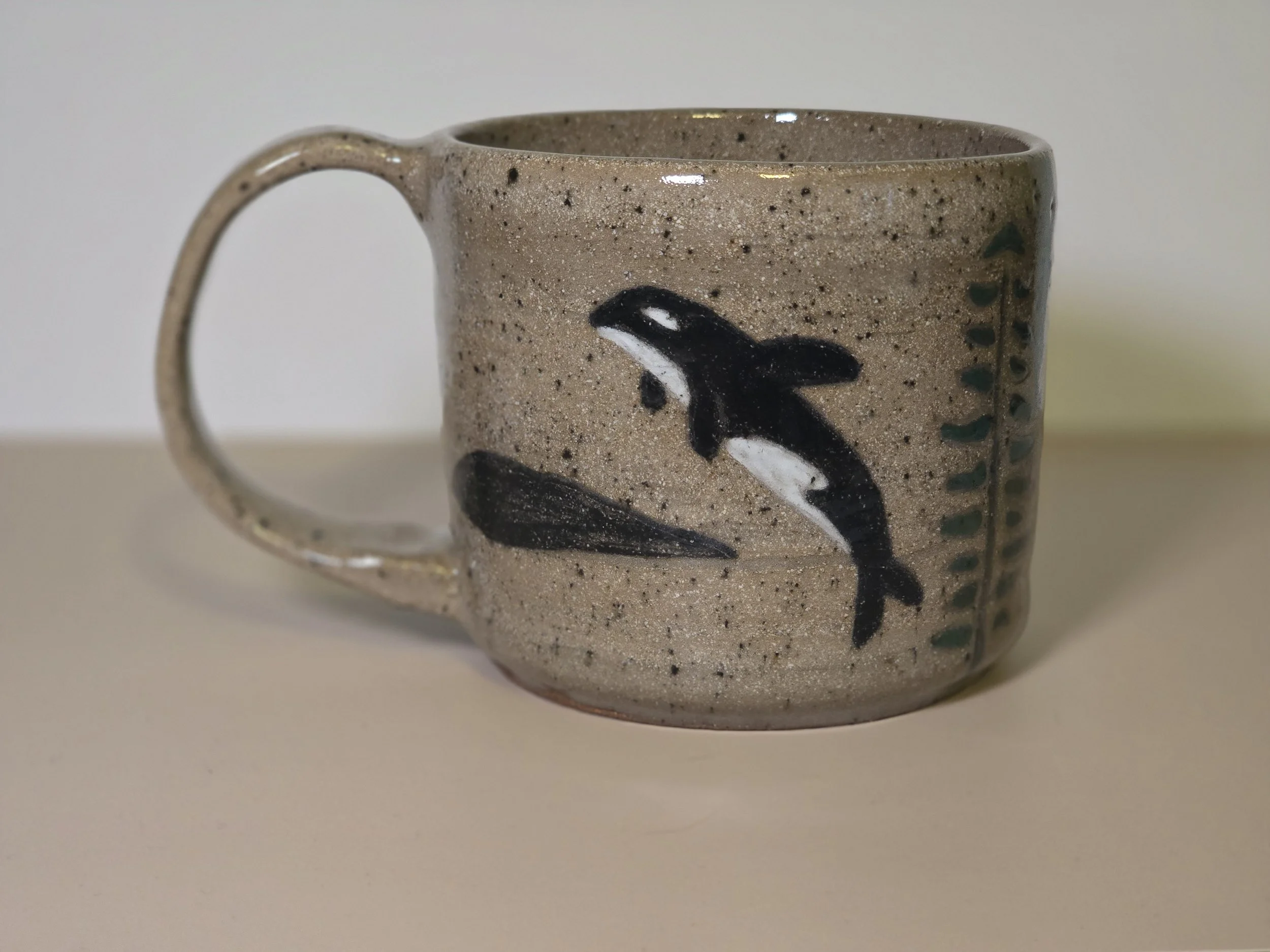 A ceramic mug featuring a painted orca whale and green seaweed design on a beige speckled background.