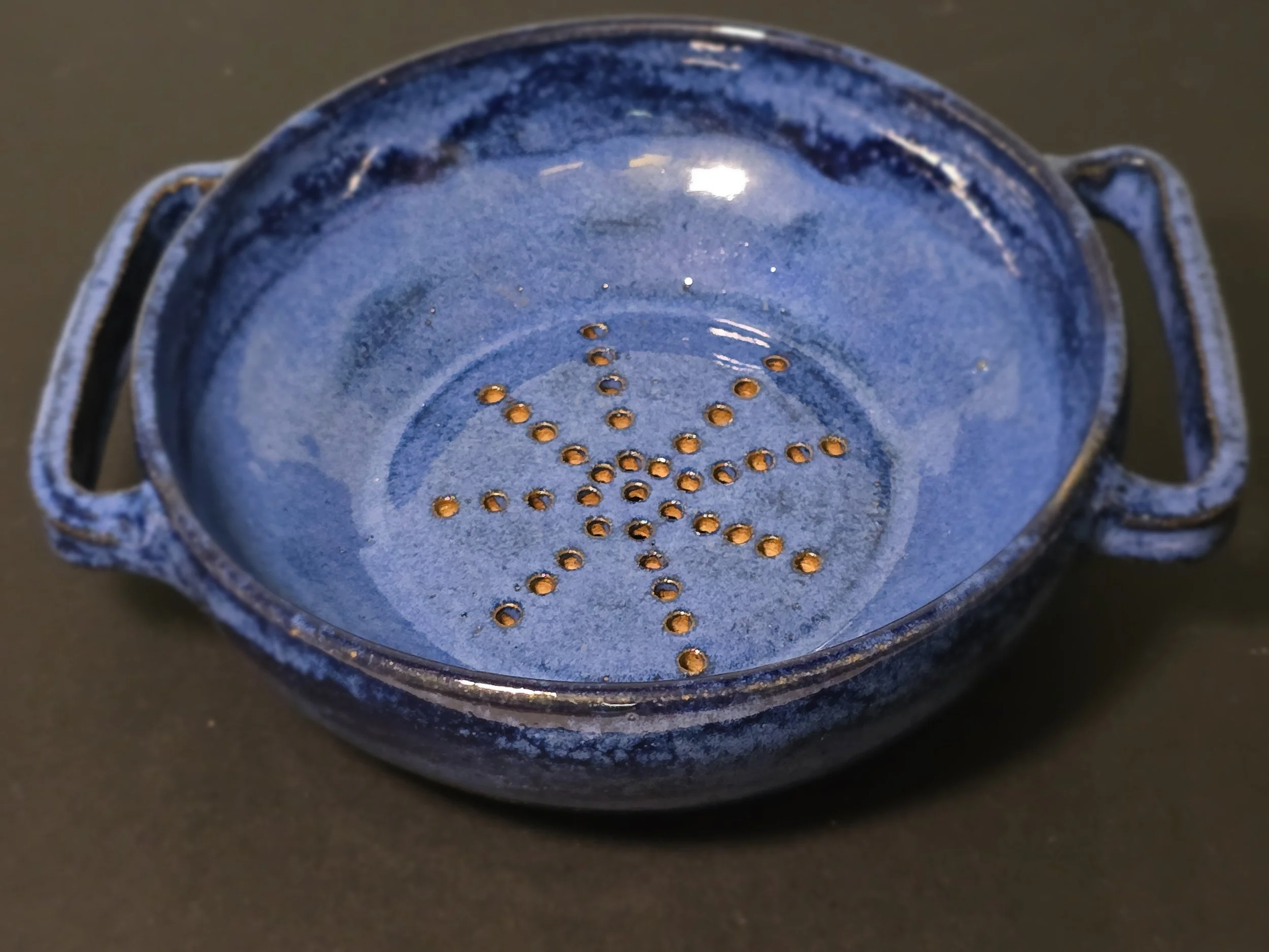 Blue ceramic strainer with small holes in the shape of a star, placed on a dark surface.