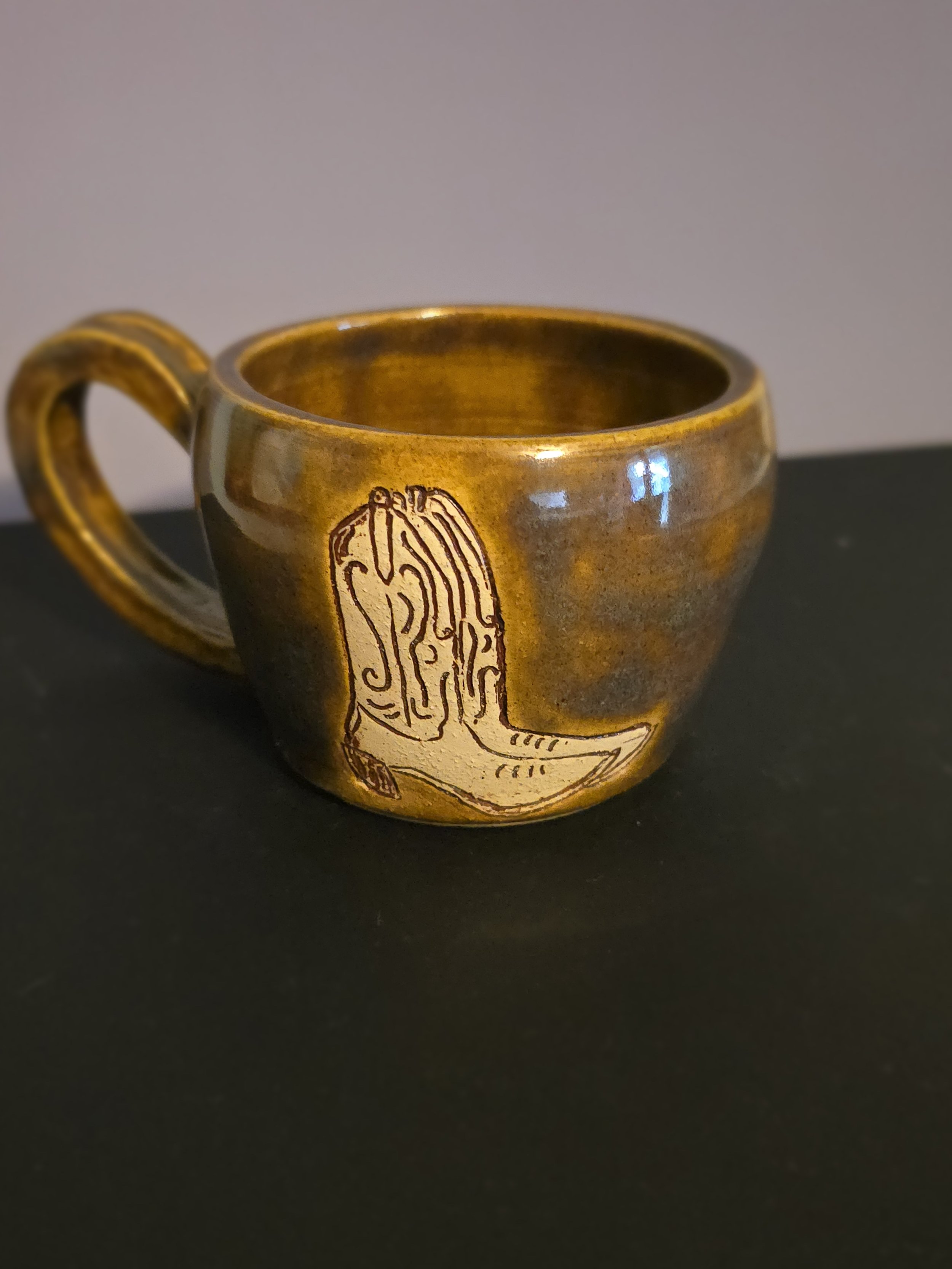 A ceramic coffee mug with a brown glaze and a design of a cowboy boot on the side, placed on a black surface against a plain background.