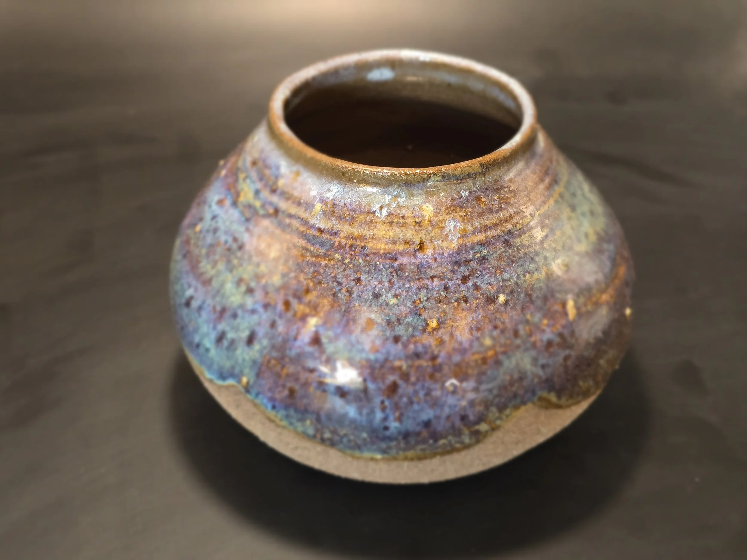 A round, rustic ceramic vase with a narrow opening, glazed in multicolored earthy tones, sitting on a dark surface.