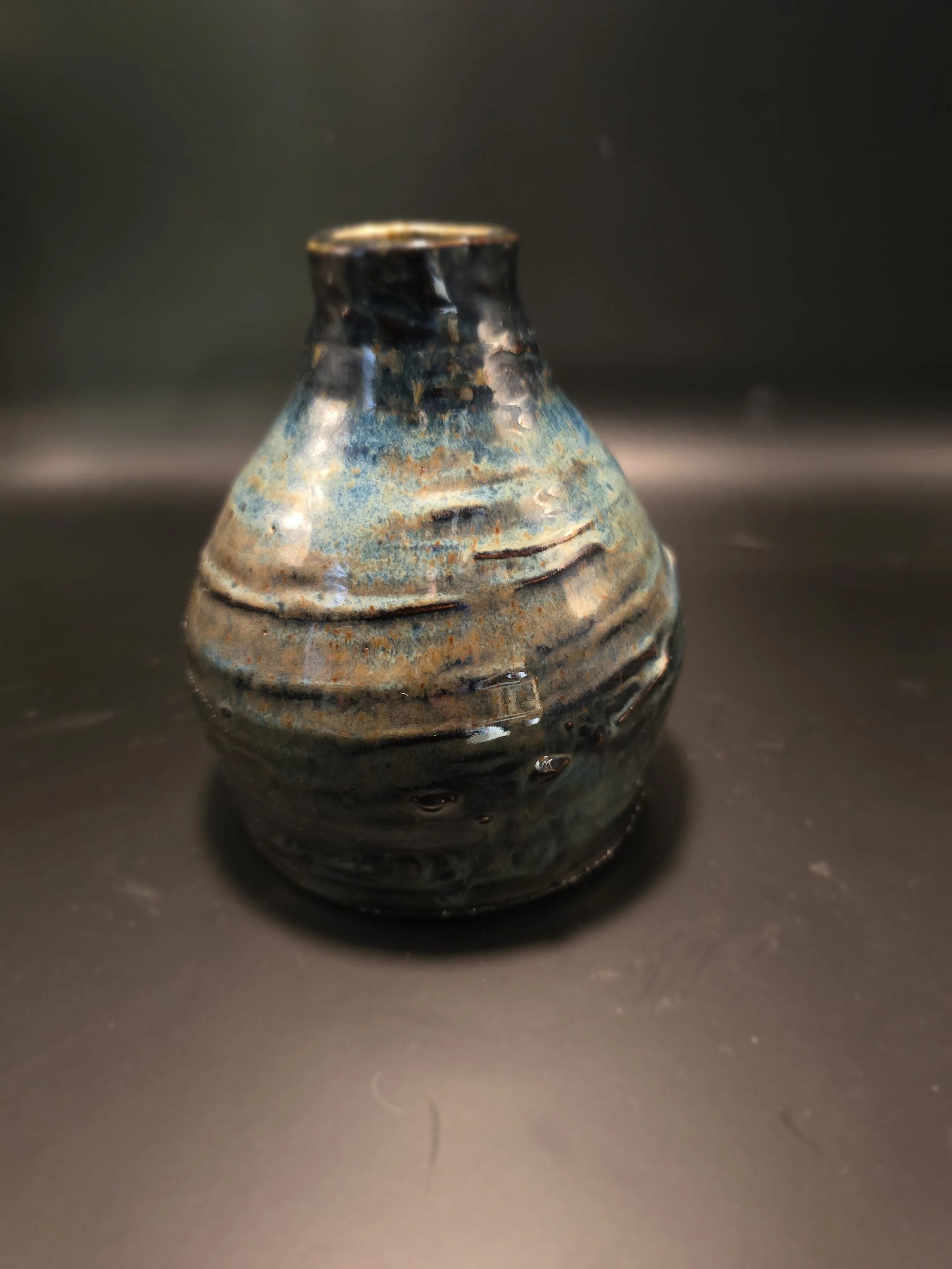 A small ceramic vase with a rounded body, narrow neck, and wide opening, featuring a textured glaze with shades of blue, black, and earthy tones.