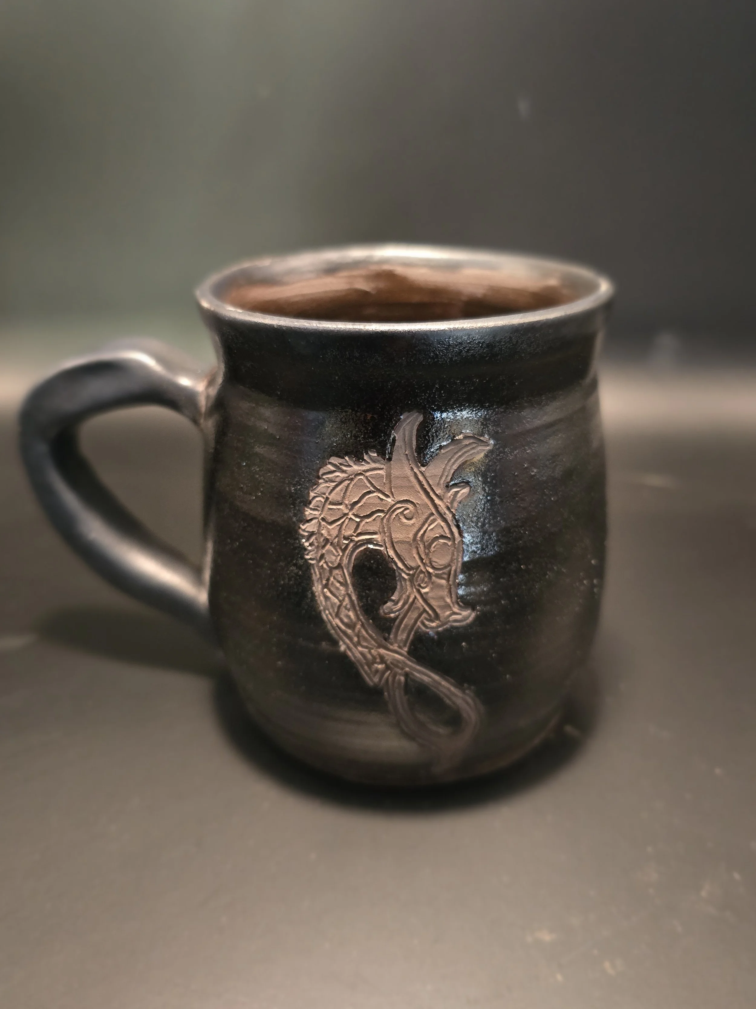 A black ceramic mug with a dragon design on the front, filled with a beverage.