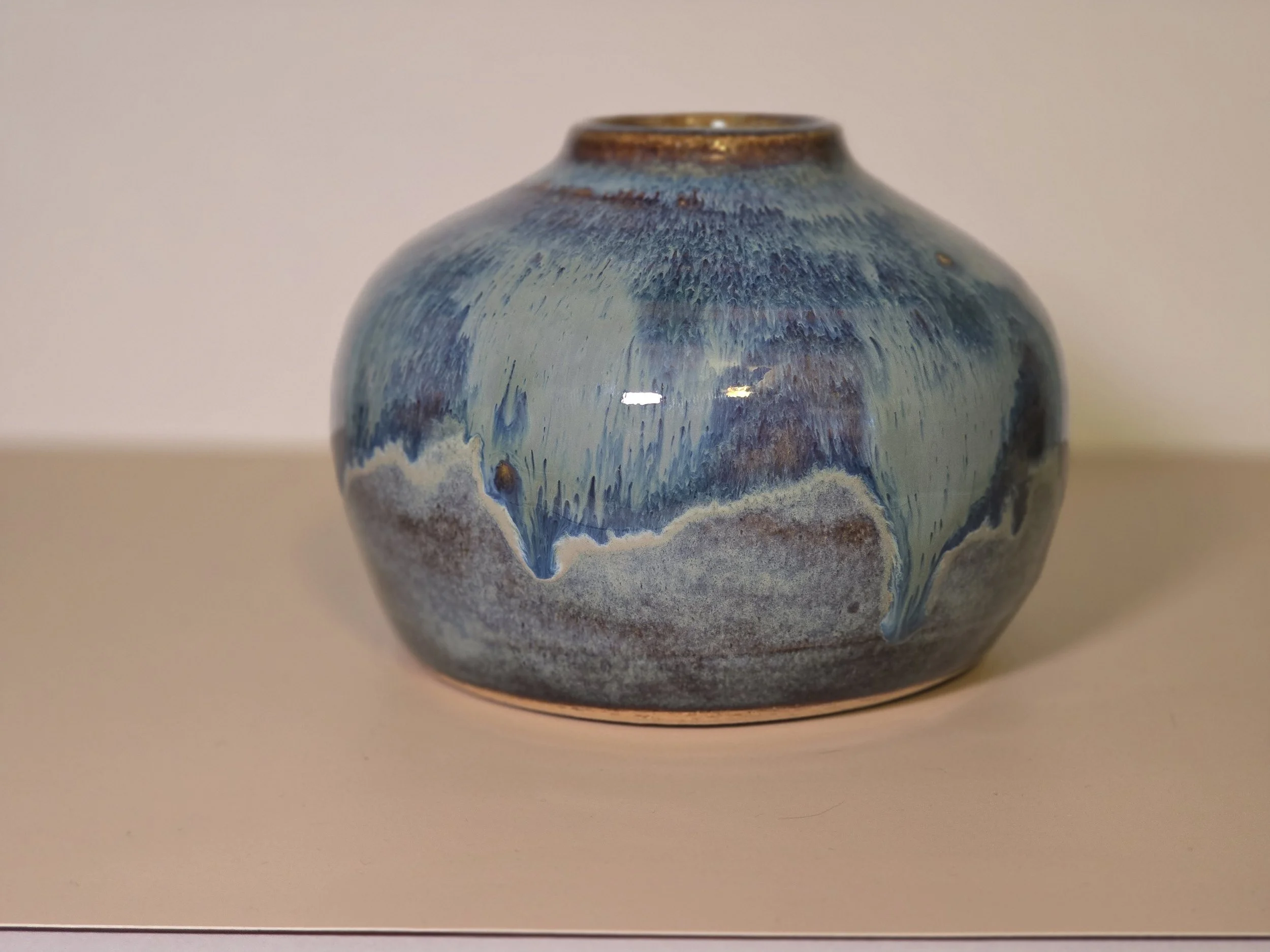 A round ceramic vase with a blue and beige glaze pattern, sitting on a white surface with a plain background.