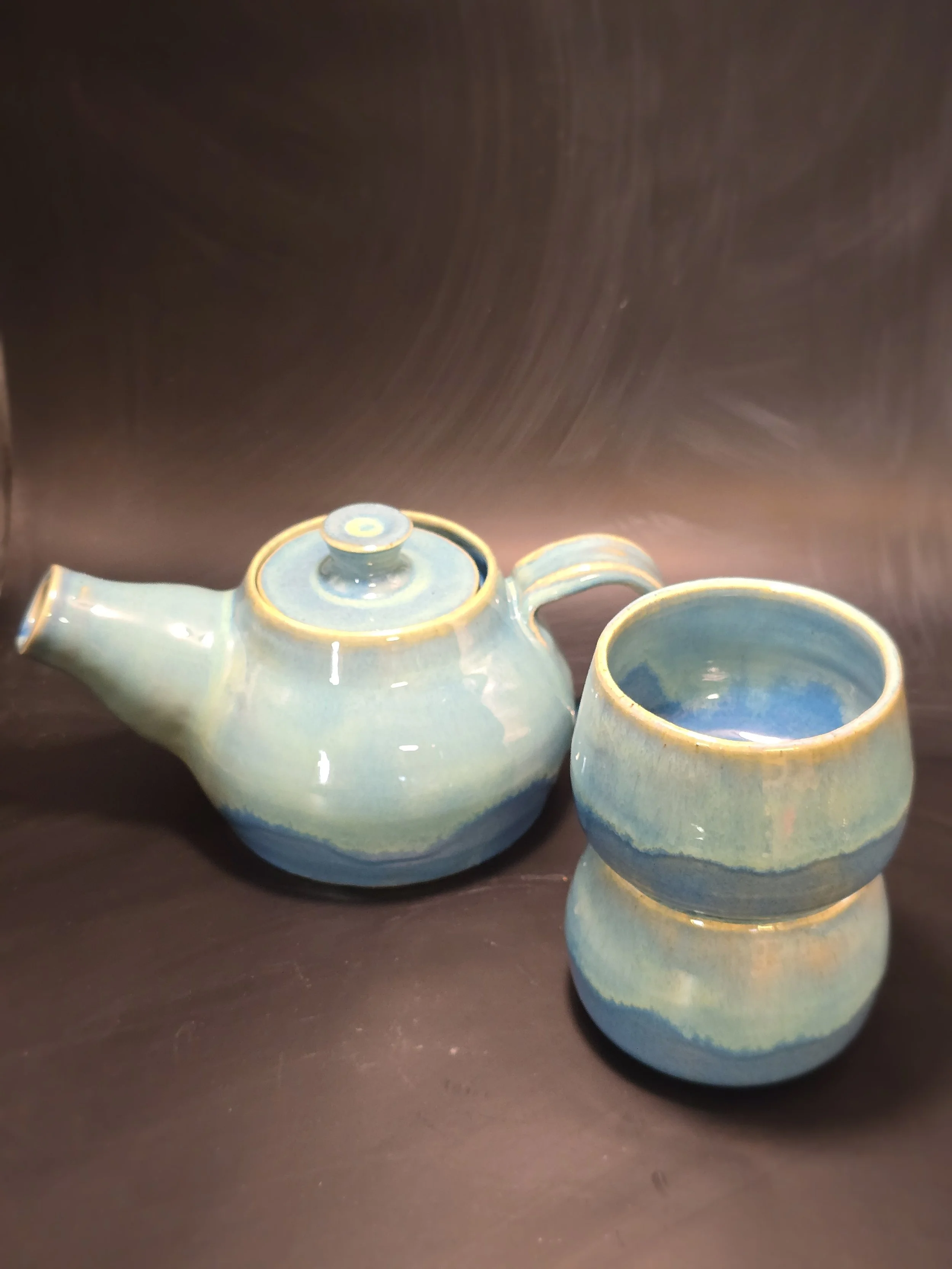 A light blue ceramic teapot with two matching cups, all featuring a darker blue band near the bottom, placed on a dark wooden surface.