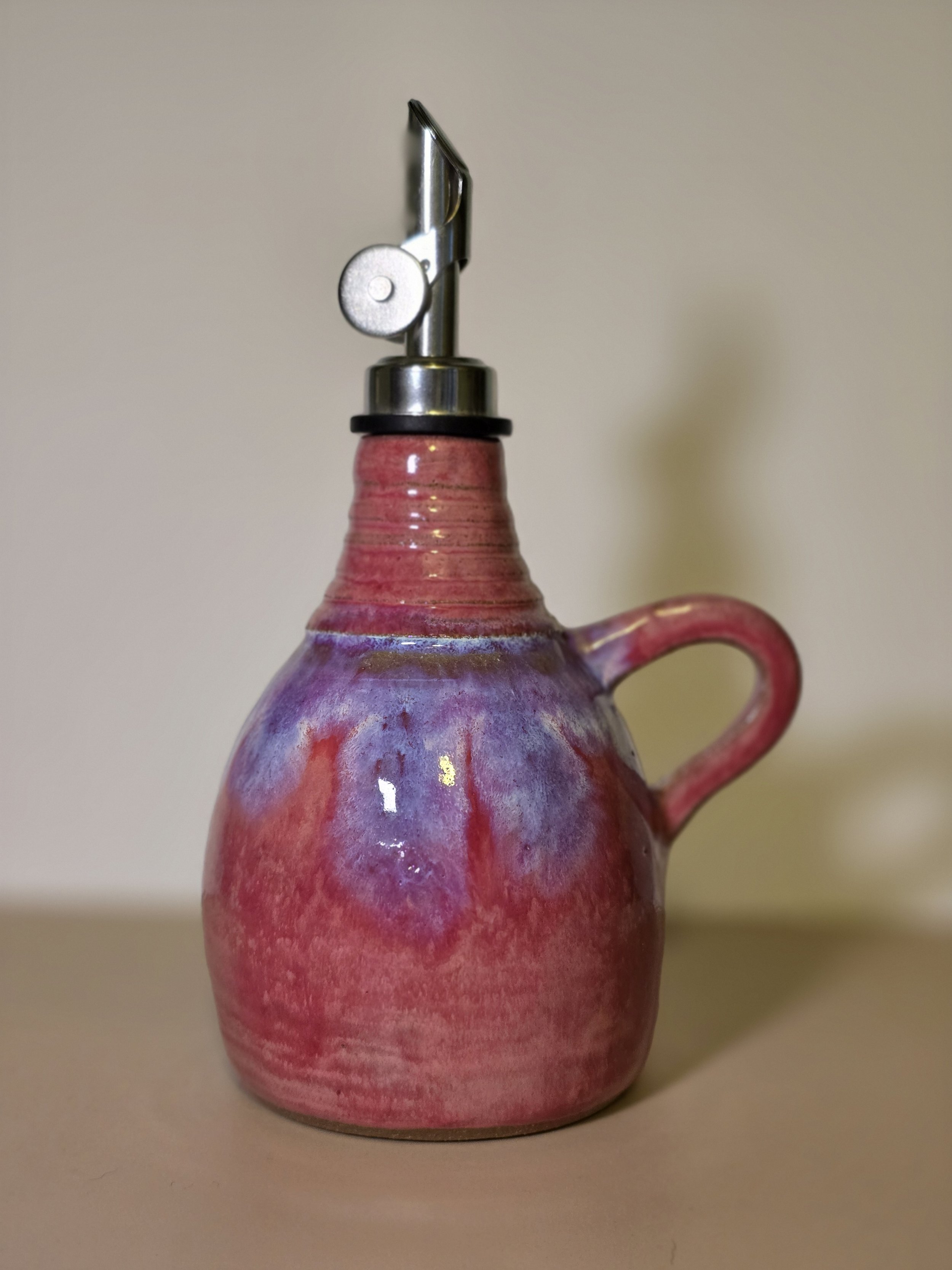 A ceramic teapot with a pink and purple glaze and a curved handle, topped with a metal pour spout and a black rubber seal, against a plain beige background.
