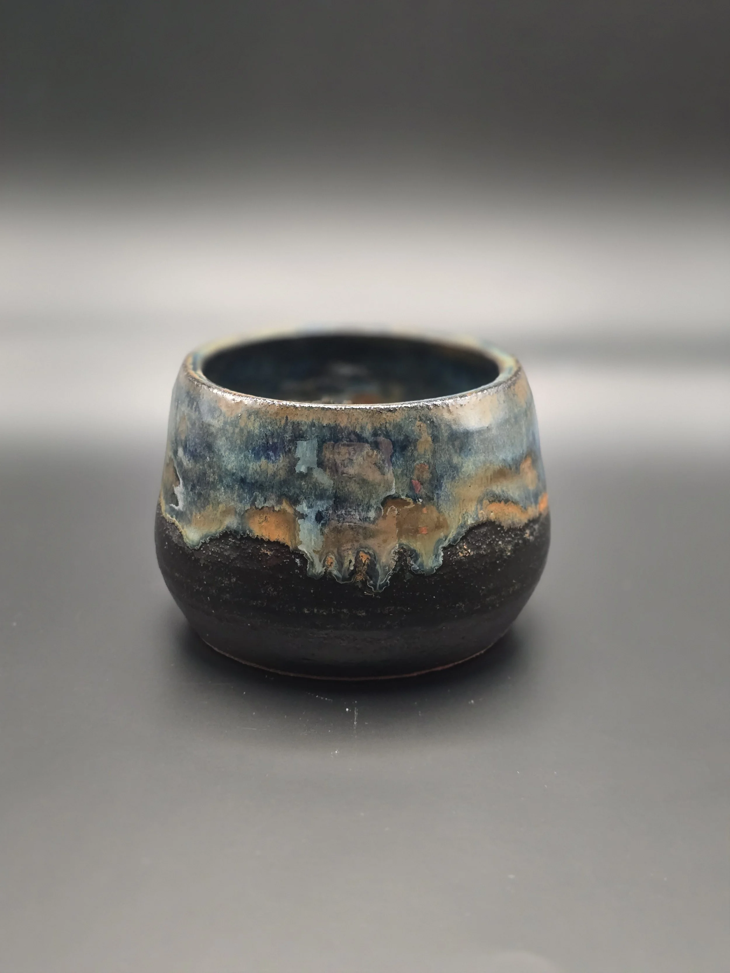 A small ceramic pot with a textured glaze and an abstract, multicolored design, on a dark gray surface with a dark gradient background.