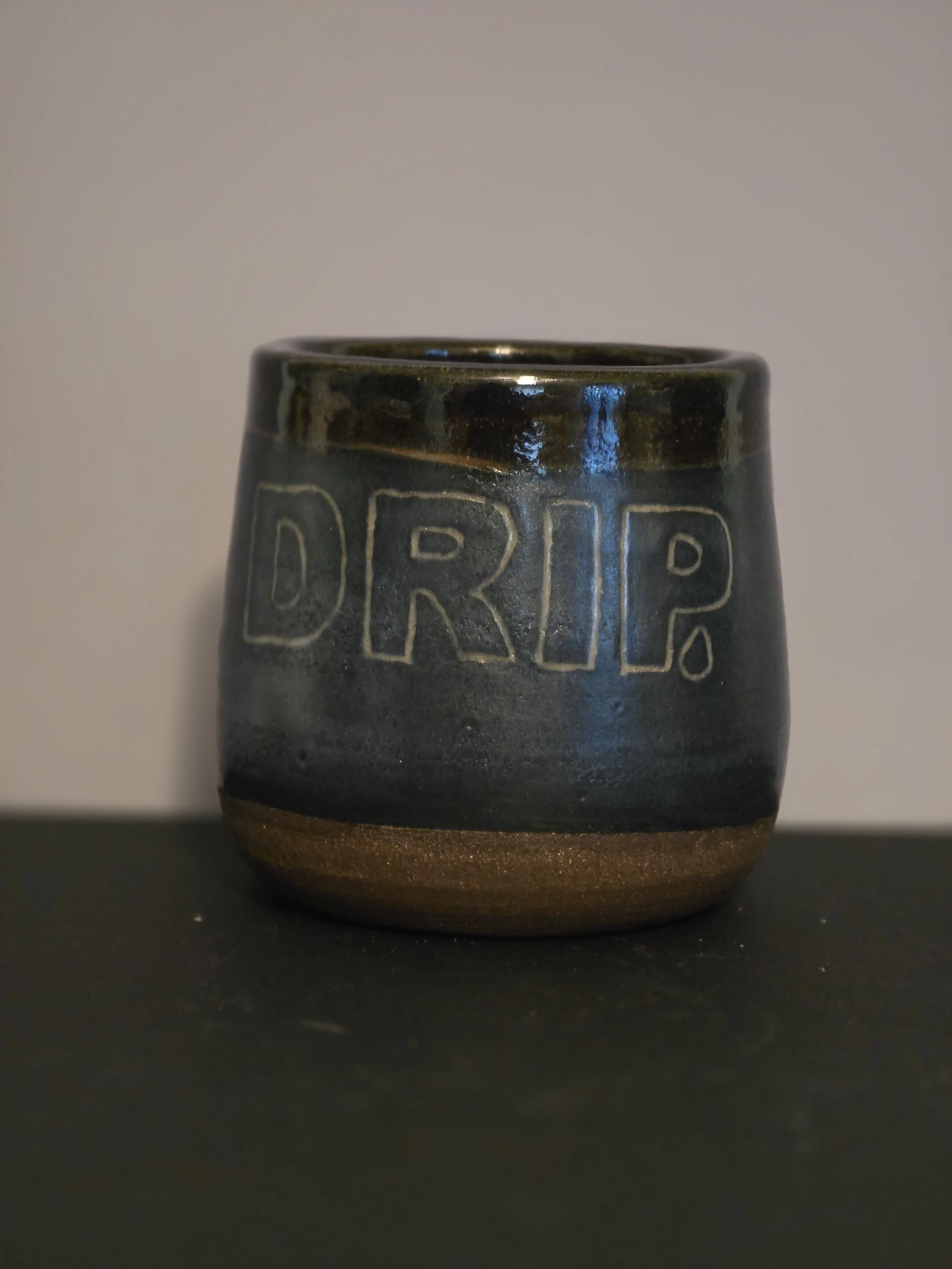 A ceramic permaculture coffee mug with the word 'DRIP' carved into its dark blue glazed surface, sitting on a black surface against a plain background.