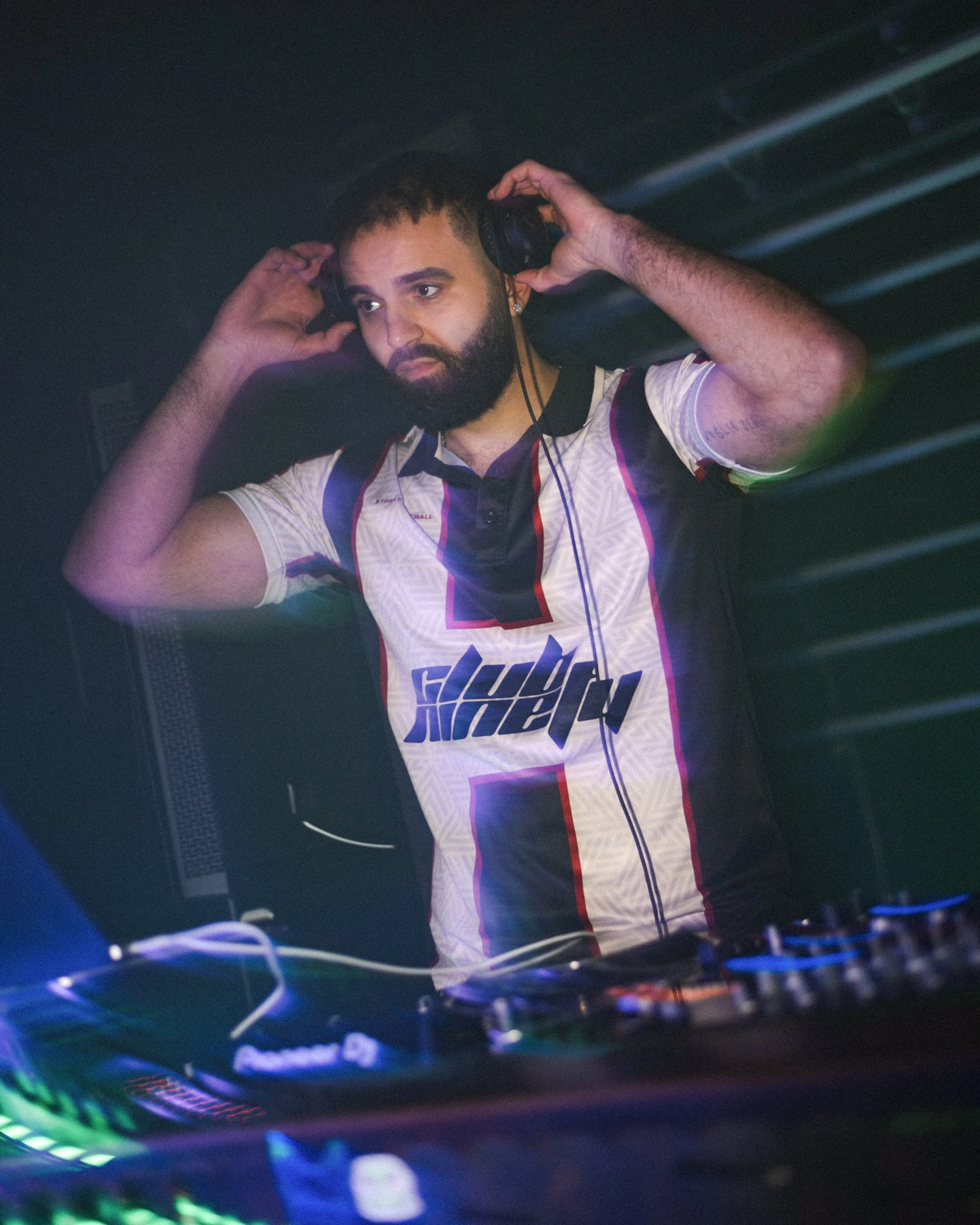 A bearded man with short dark hair wearing a racing jersey with a colorful design and the word 'FIGHT' on it, adjusting large black headphones in a dimly lit room with LED lights and DJ equipment.
