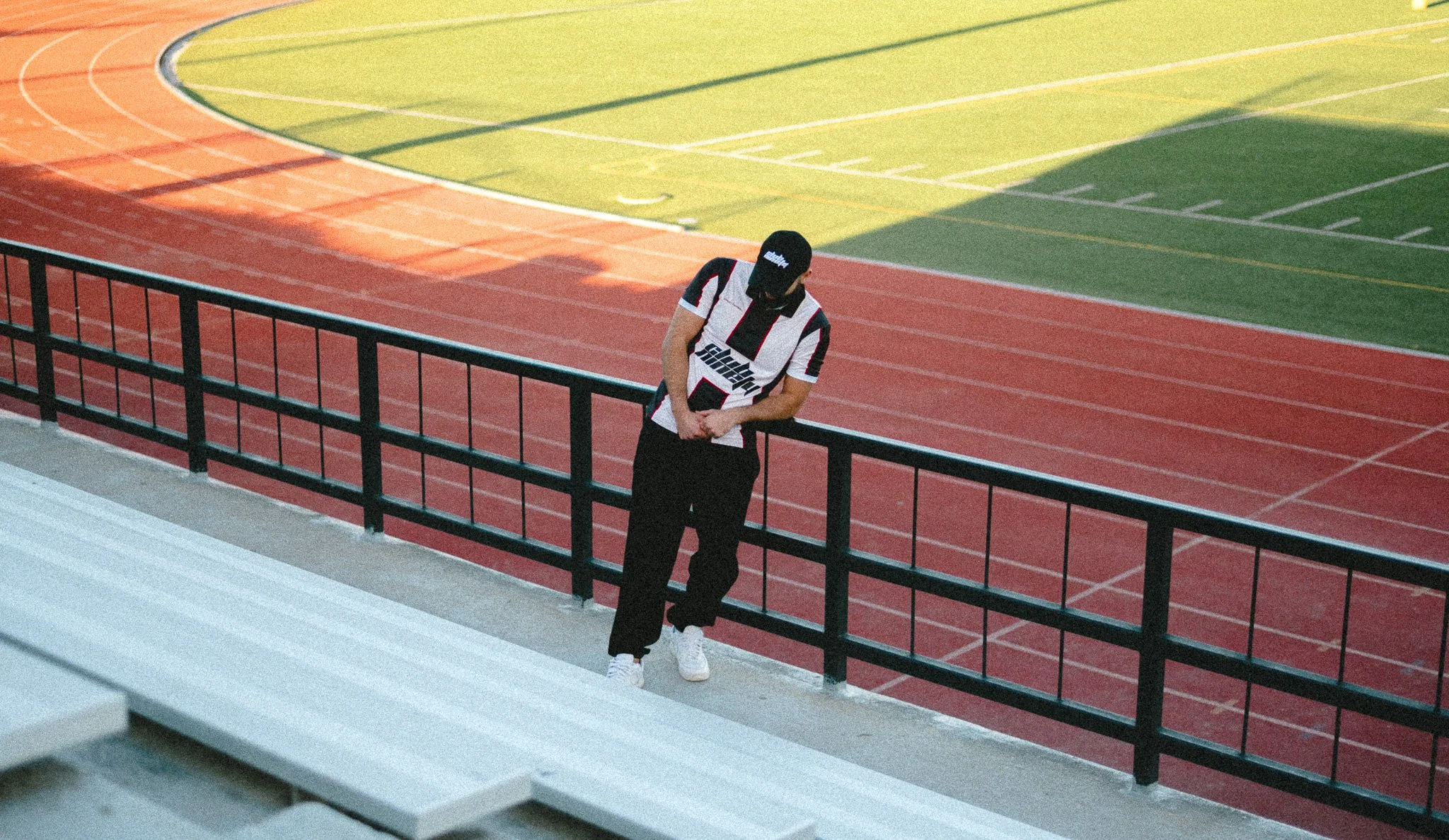A person wearing a black cap, a white and black sports jersey, black pants, and white shoes stands on a raised platform next to a sports field. The field has a red running track and green grass with white painted lines. The person is looking down and holding their hands in front of them, with their head slightly bowed.