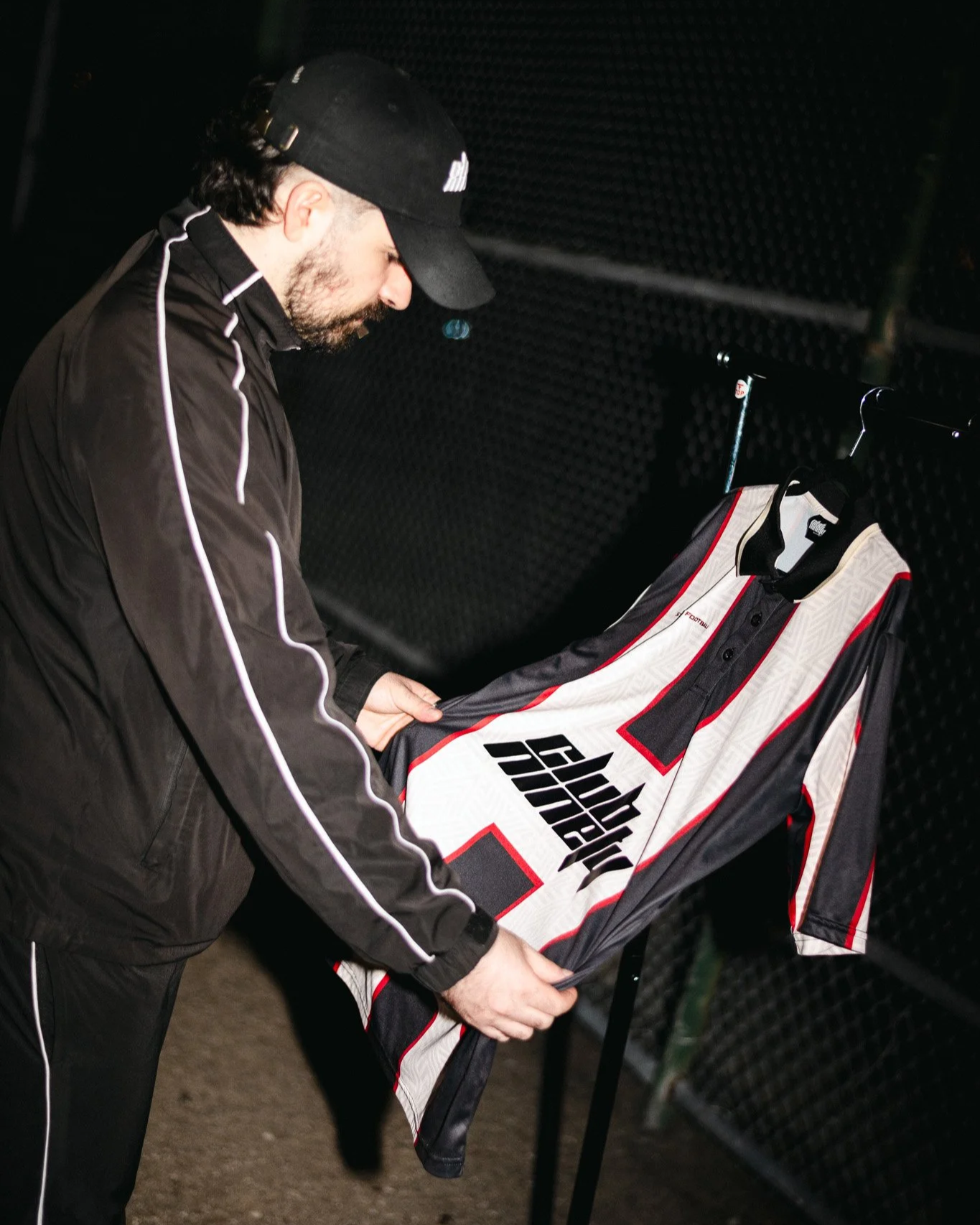 A man wearing a black jacket and black cap is hanging a hockey jersey on a clothing rack. The jersey is white with black and red accents and displays the text 'Hockey' on the front. The background features a dark, textured wall and fencing.