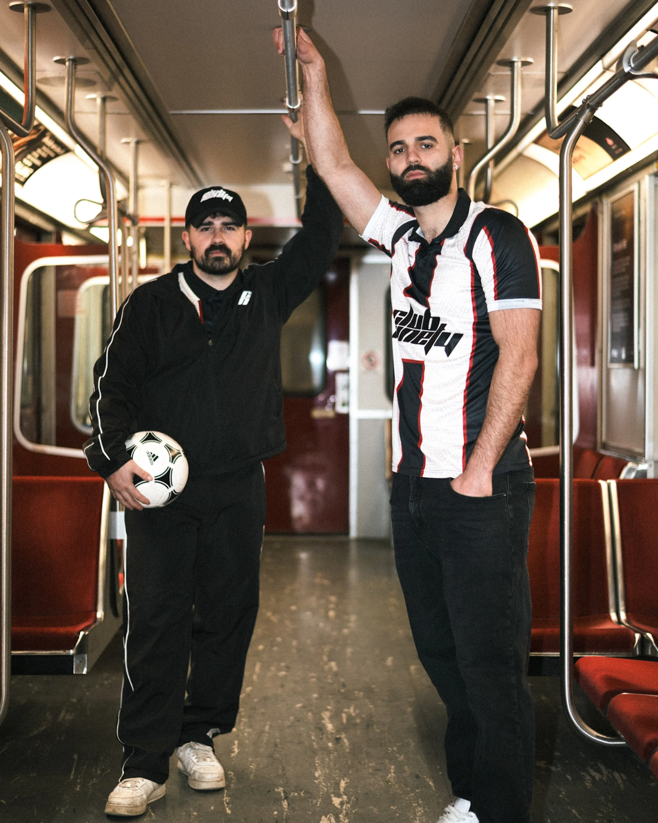 Two men standing inside a subway train. The man on the left is wearing a black hoodie, black pants, white sneakers, a black cap, and is holding a soccer ball. The man on the right is wearing a black, red, and white sports jersey, black pants, and white sneakers. They are both looking at the camera, with the man on the right holding onto a horizontal handrail above.