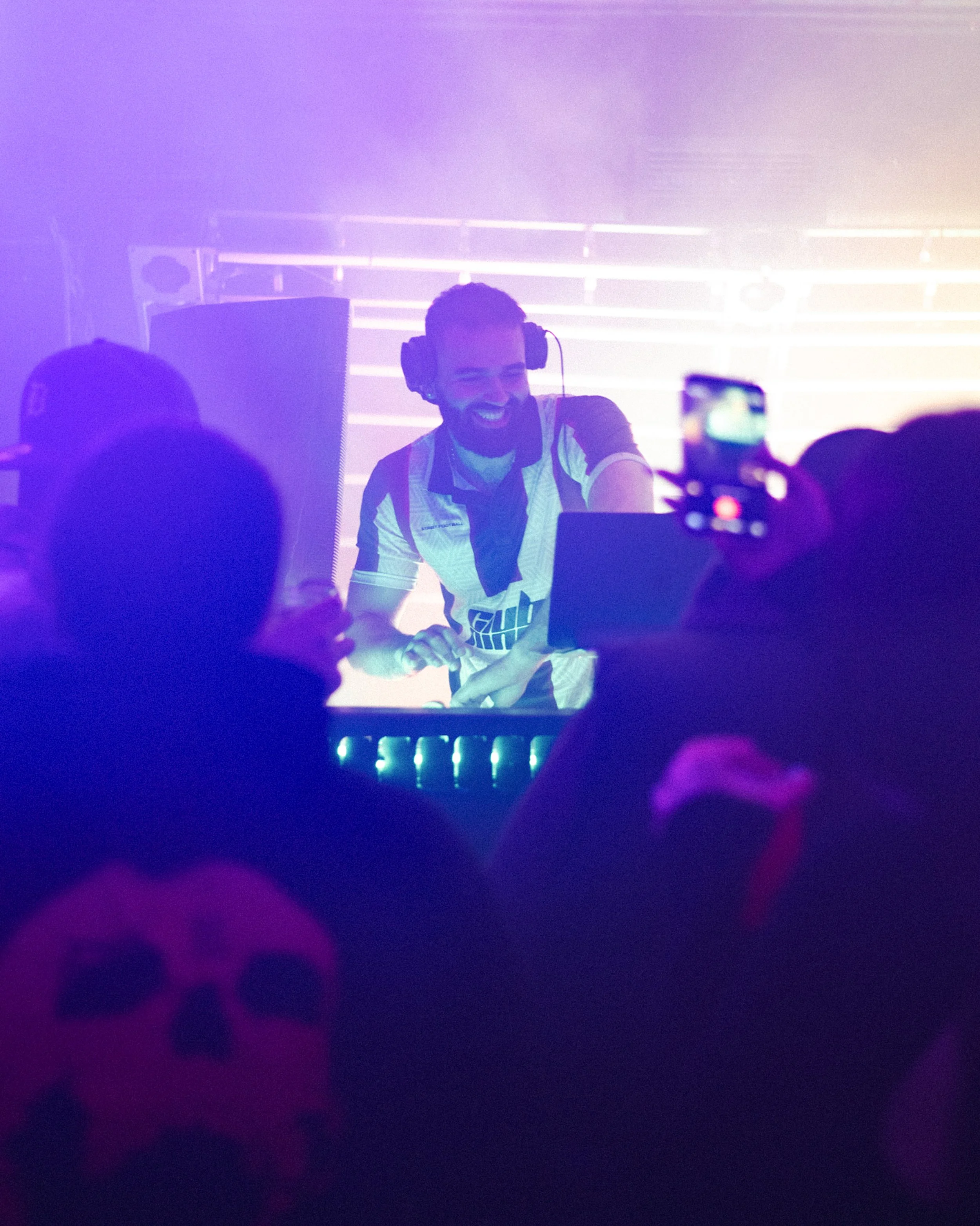 Person with a beard wearing headphones and a sports jersey performing as a DJ at a music event with purple and pink lighting.