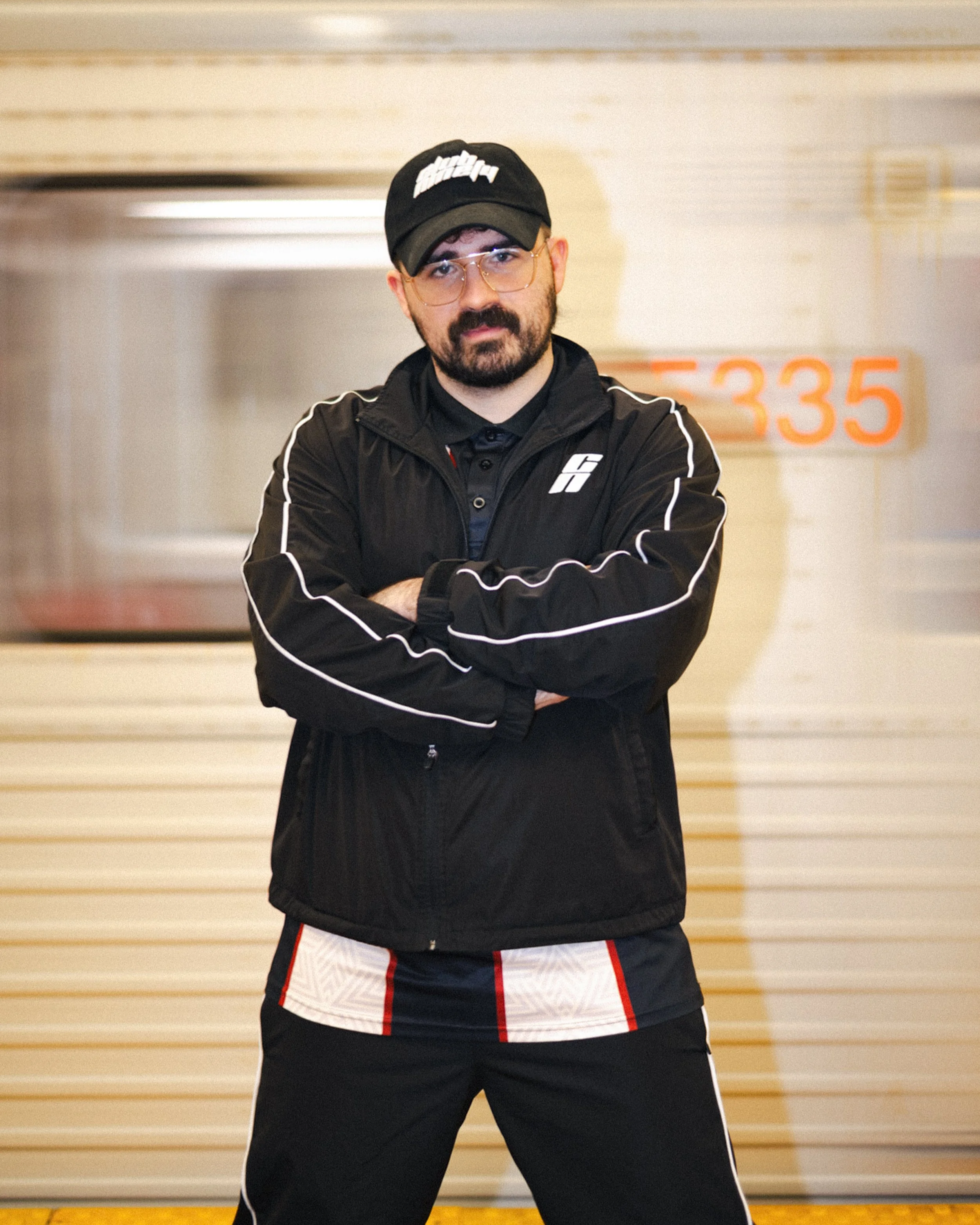 A man in a black sports jacket and shorts standing with arms crossed in an indoor sports facility, with a background of a wooden wall and a sign with numbers '5 33 5' in orange.