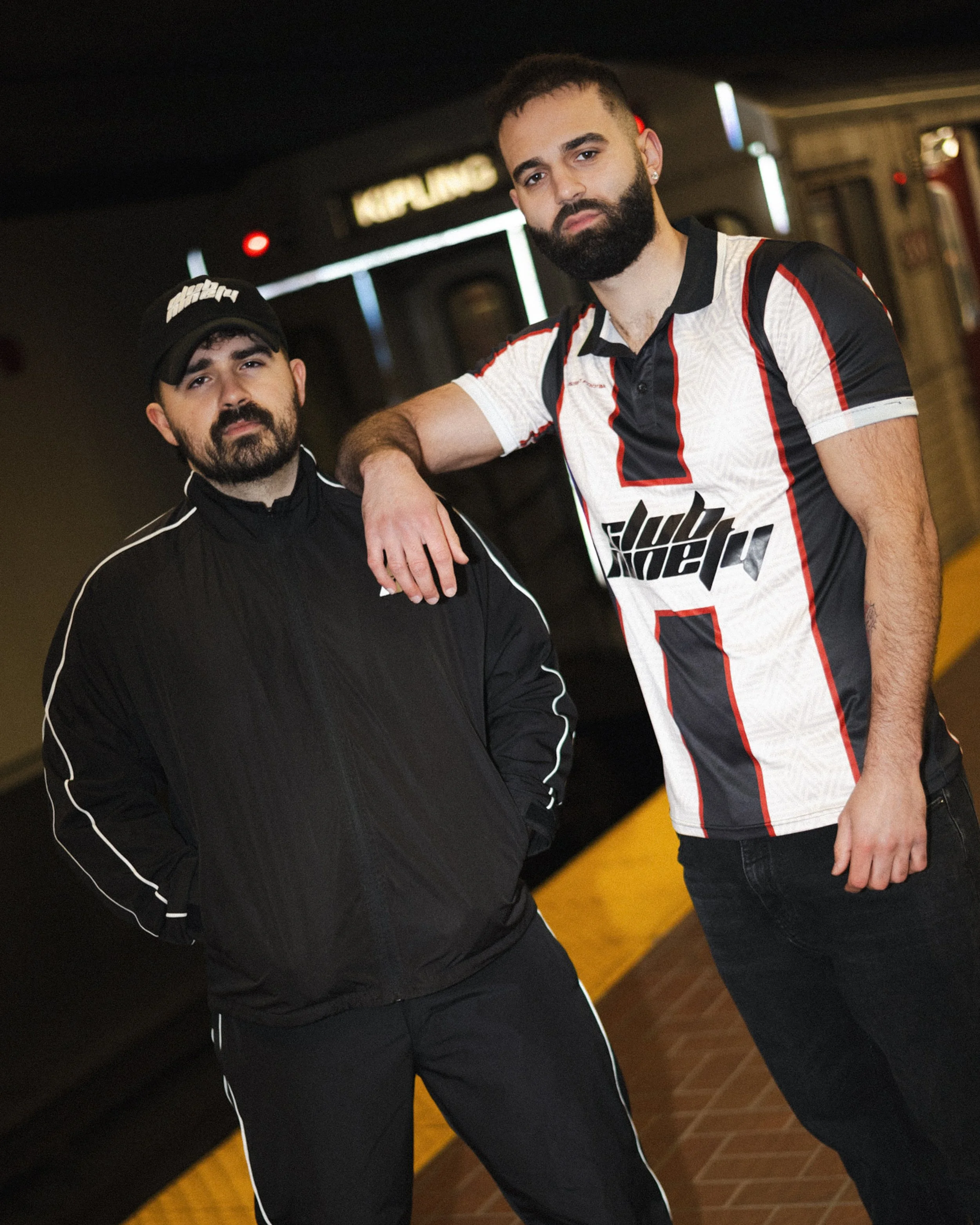 Two men stand together in an indoor setting, with one resting his arm on the other's shoulder. The man on the right wears a white, black, and red sports jersey, has a beard, and short dark hair. The man on the left wears a black jacket with white piping, a black cap, and has a beard and short dark hair. Both have serious expressions, and the background includes a sign with blurred text and dim lighting.