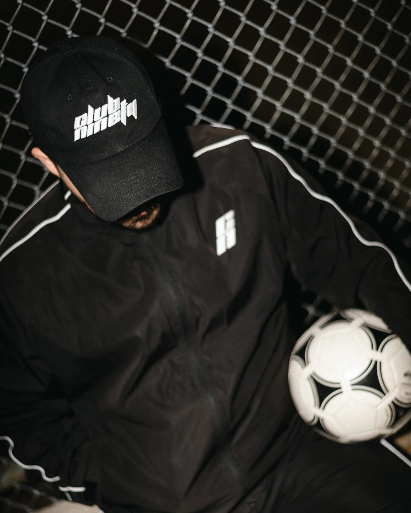 A man wearing a black Adidas jacket, black cap, and sitting on a black metal grid bench or wall, with a white soccer ball on the ground next to him.