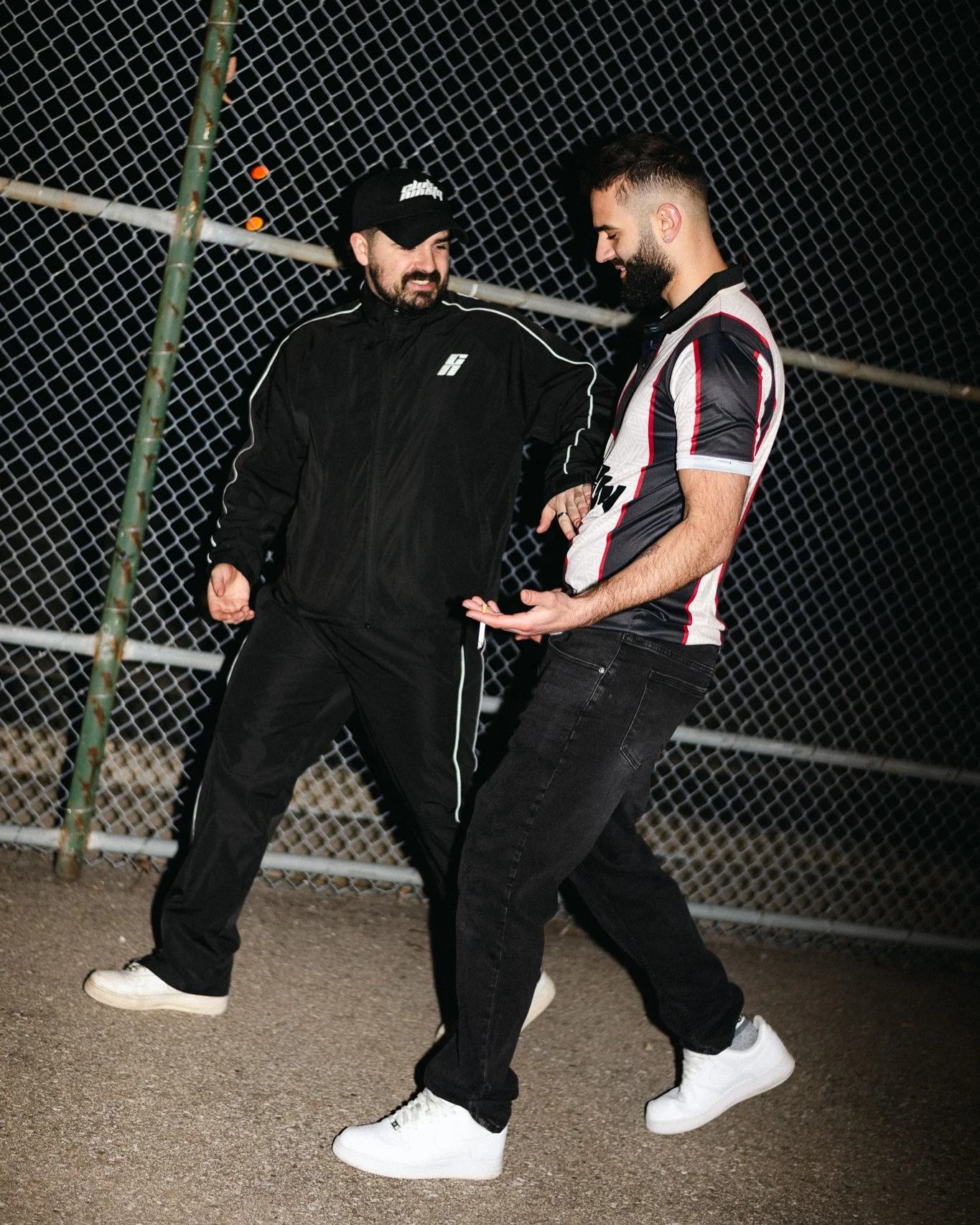 Two men dancing in front of a chain-link fence at night. One man is wearing a black tracksuit and cap, the other is in a black and white striped shirt and black jeans.