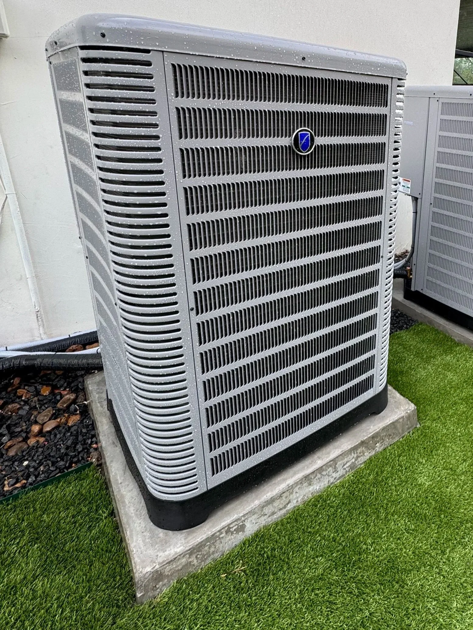 An air conditioning or heat pump unit installed outdoors on a concrete pad, surrounded by artificial grass and gravel.