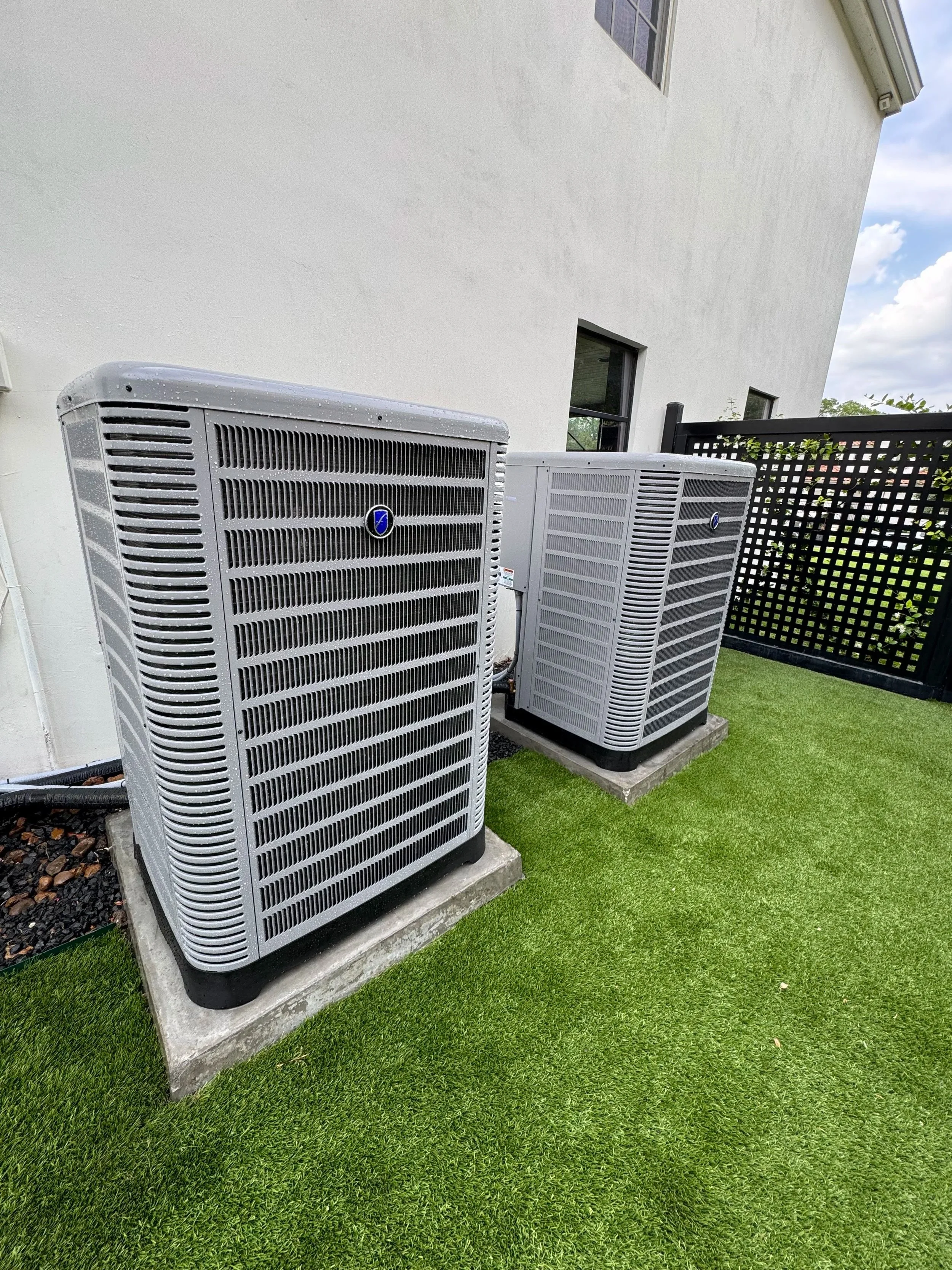 Two large outdoor air conditioning units installed on concrete slabs next to a white house with windows, surrounded by a black lattice fence and green grass.