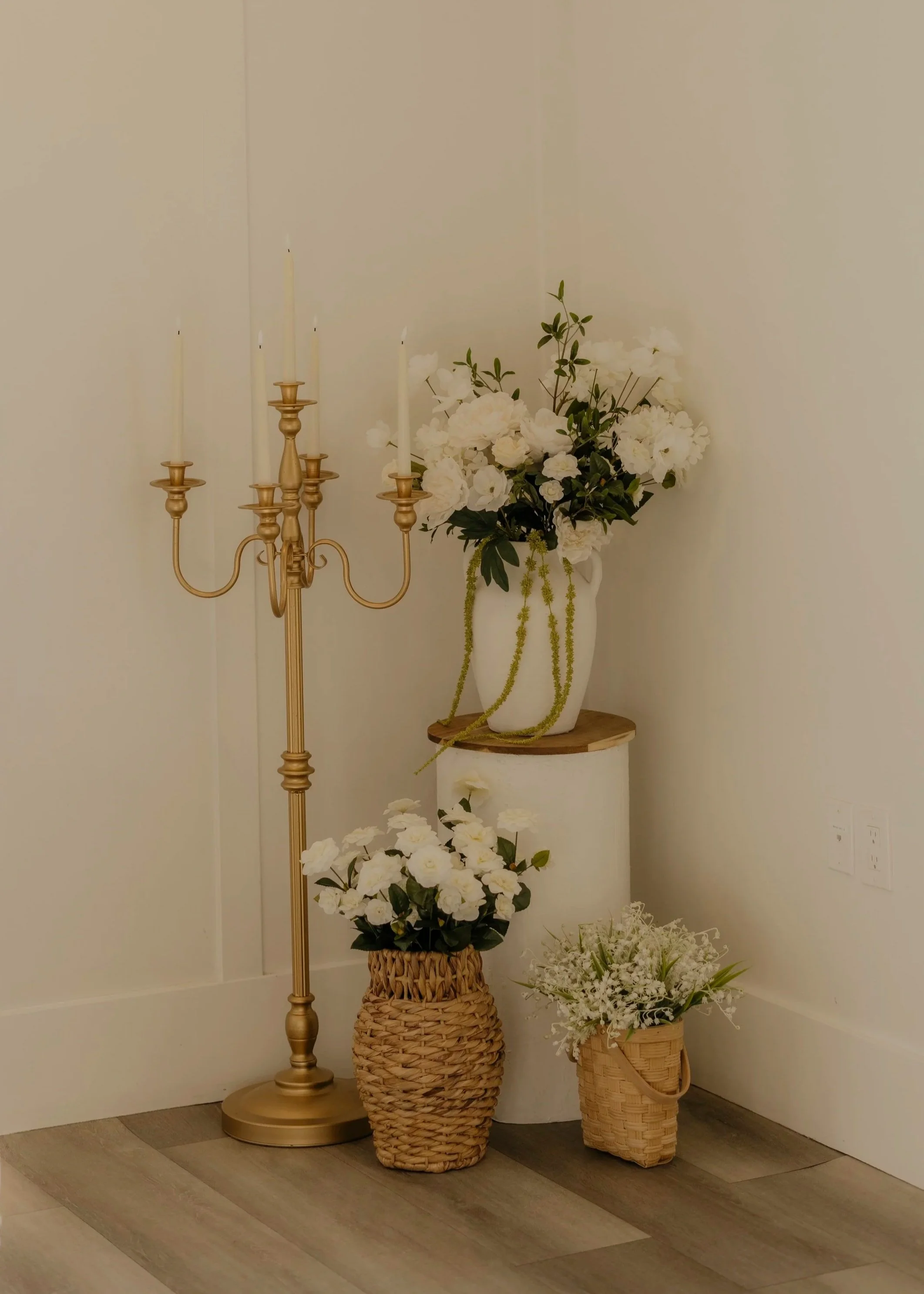 A corner with a gold candelabra, white floral arrangements in vases, and woven baskets, decorated in neutral tones.