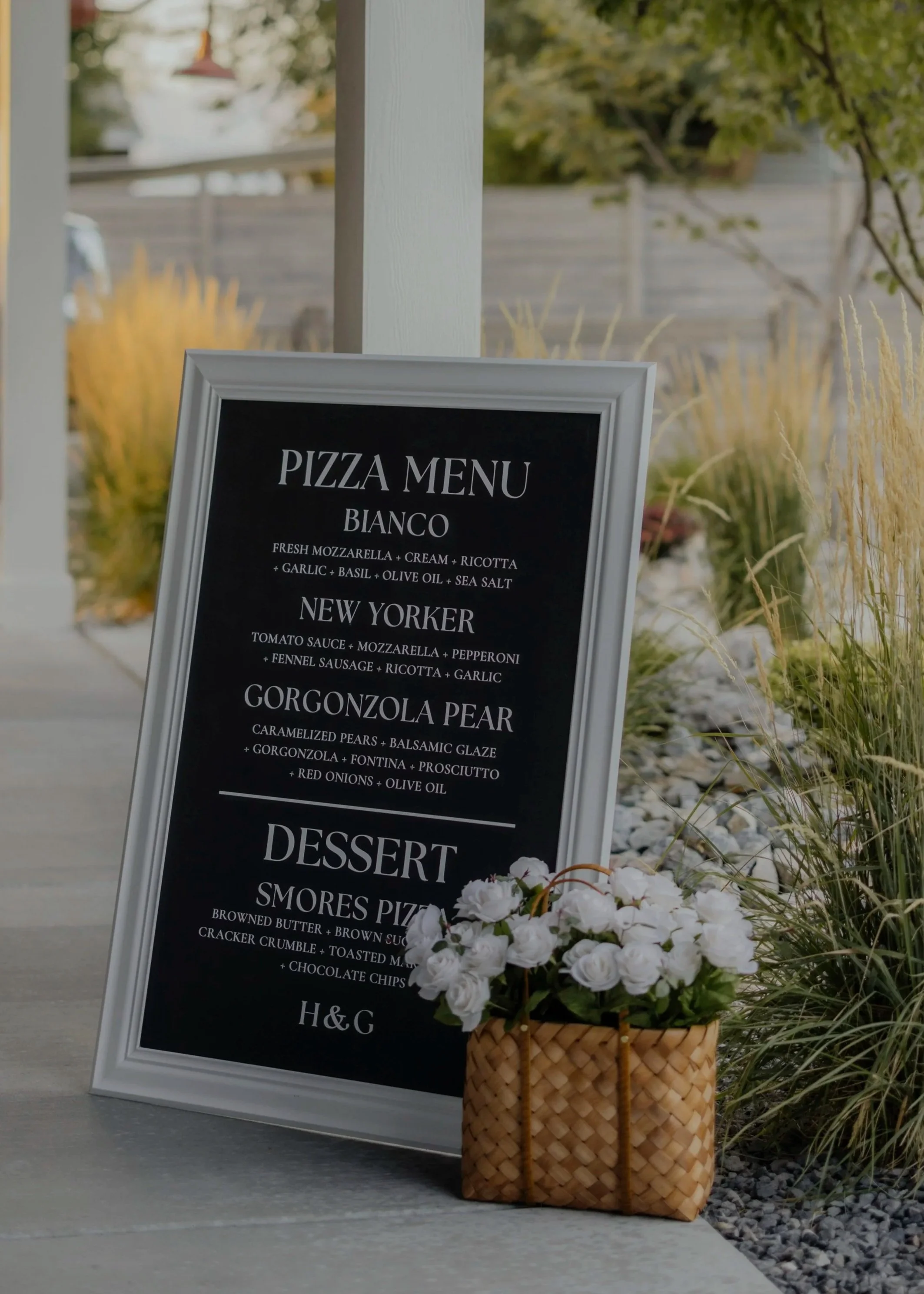 Black chalkboard sign with white framing displaying a pizza menu, placed on a ledge outdoors, next to a basket of white flowers, with plants and a garden in the background.