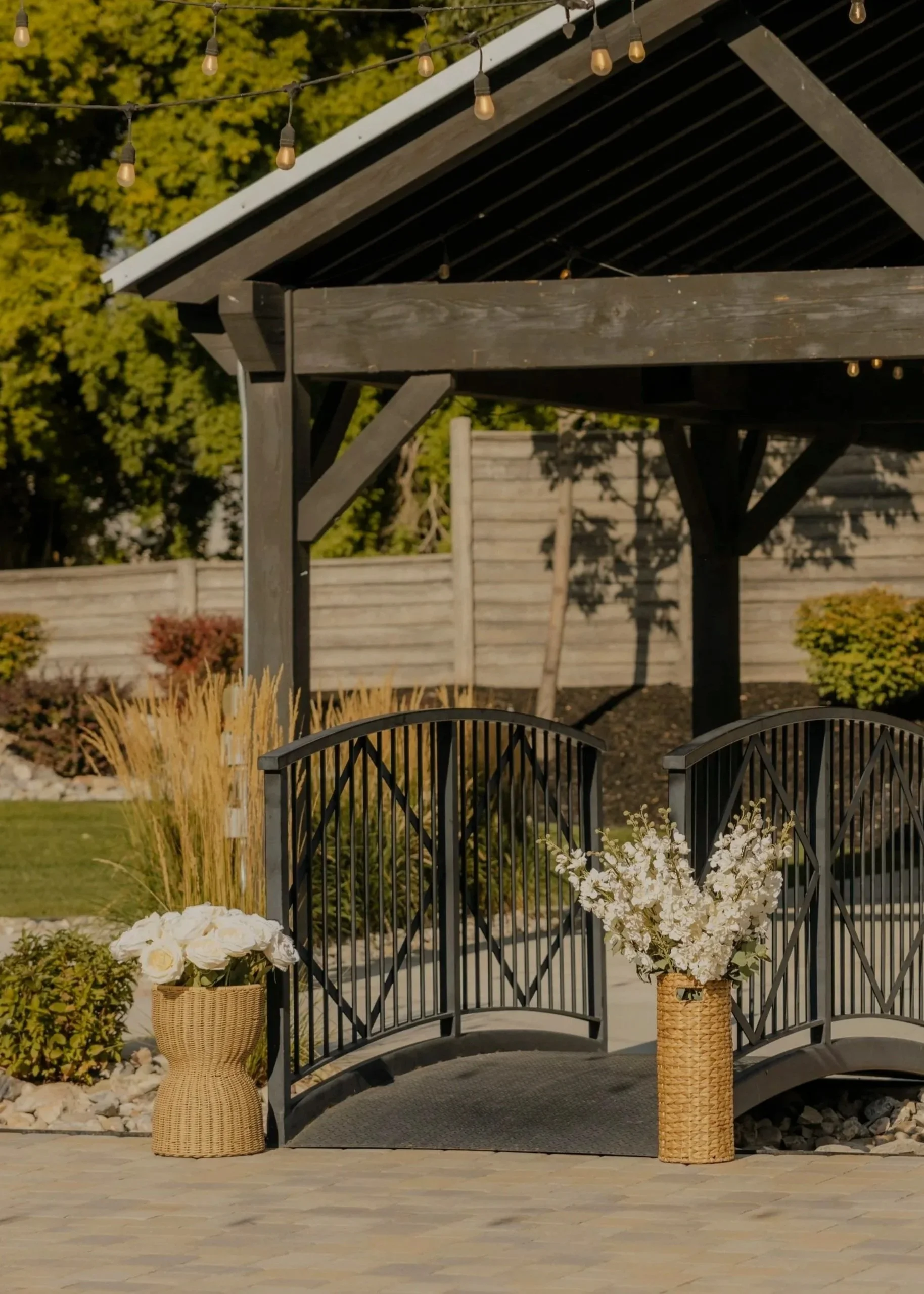 Decorative garden bridge with black railings and two baskets of white flowers on each side, set in a landscaped backyard with a wooden pergola and fencing in the background.