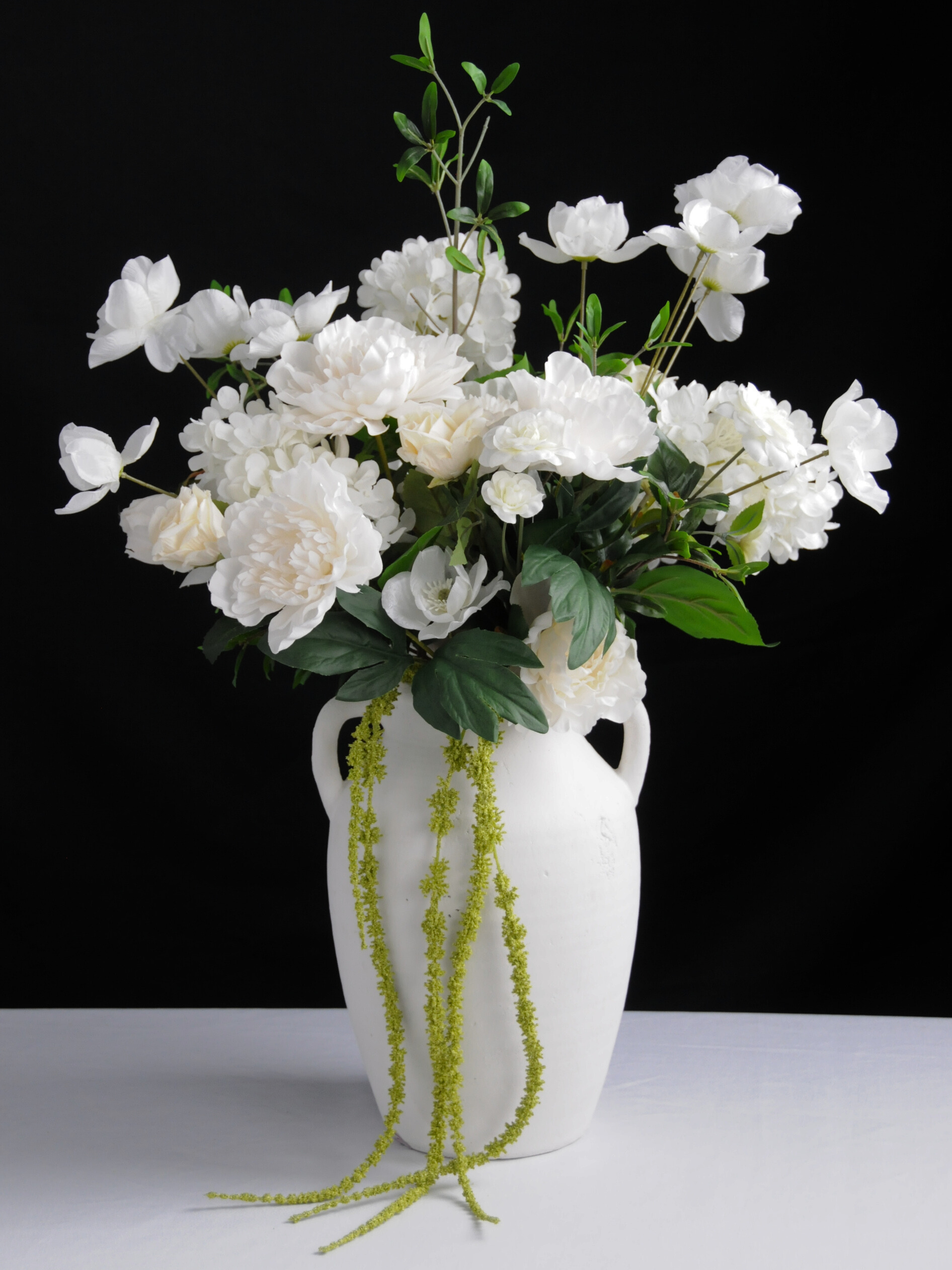 White floral arrangement with various white flowers and green leaves in a white vase, set against a black background.