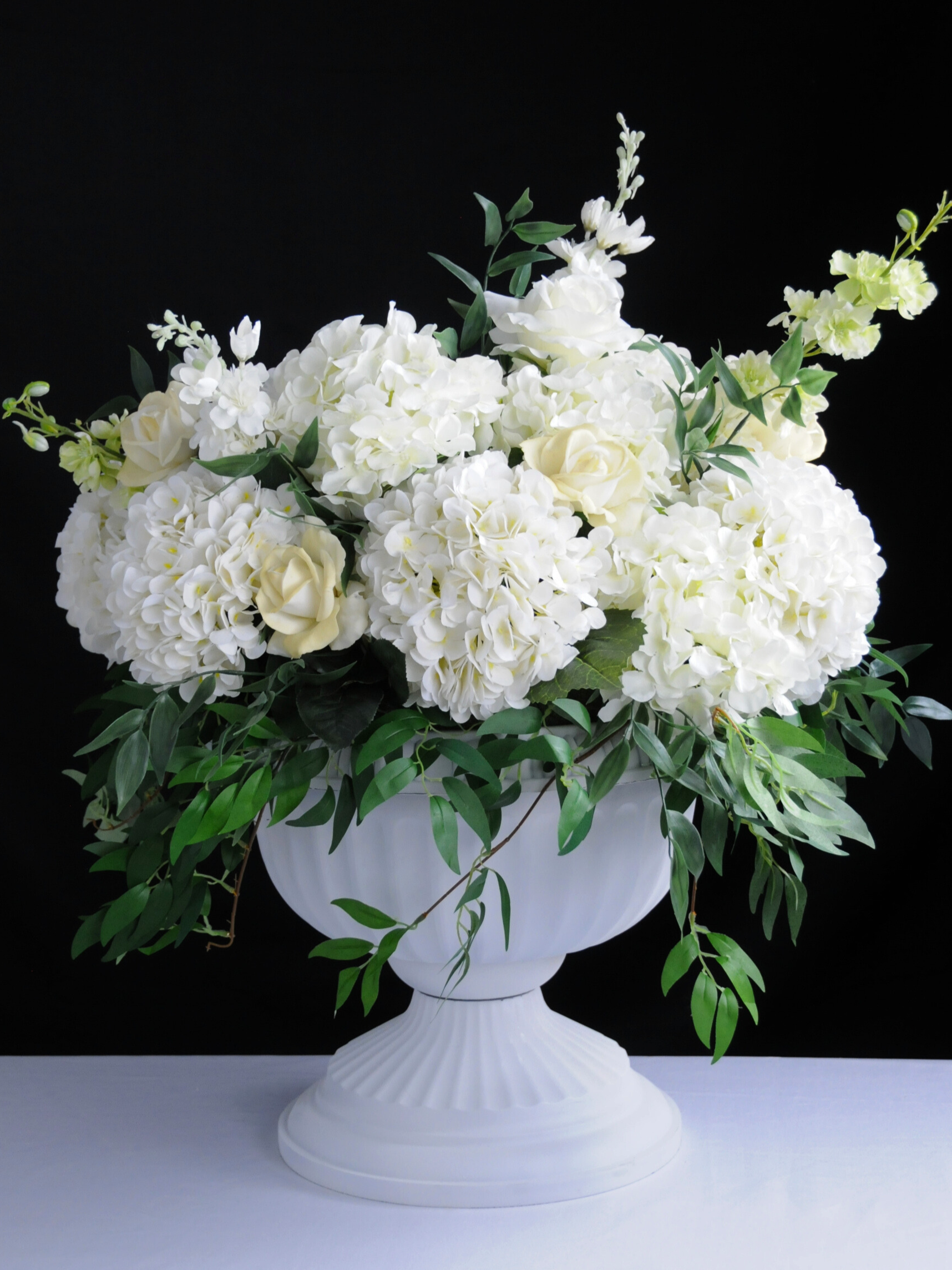A white flower arrangement in a white vase with white hydrangeas, roses, and greenery against a black background.