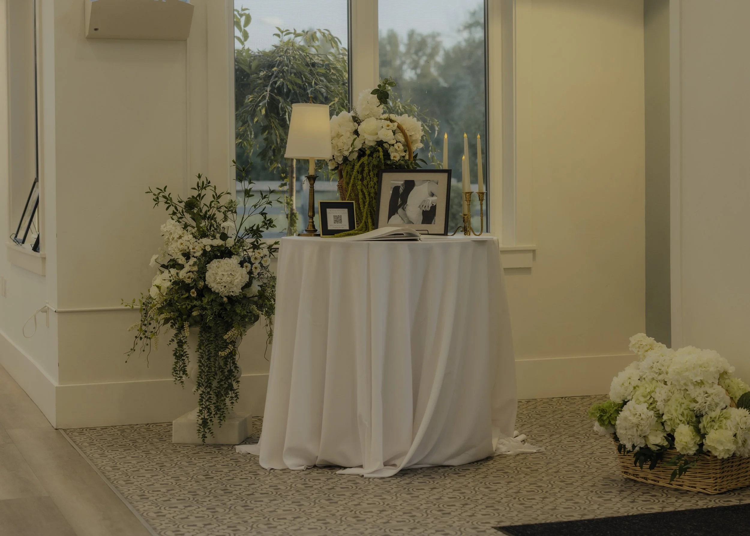 Funeral memorial display with a framed photograph, white flowers, candles, and a table draped with a white cloth, located inside a room near a window.