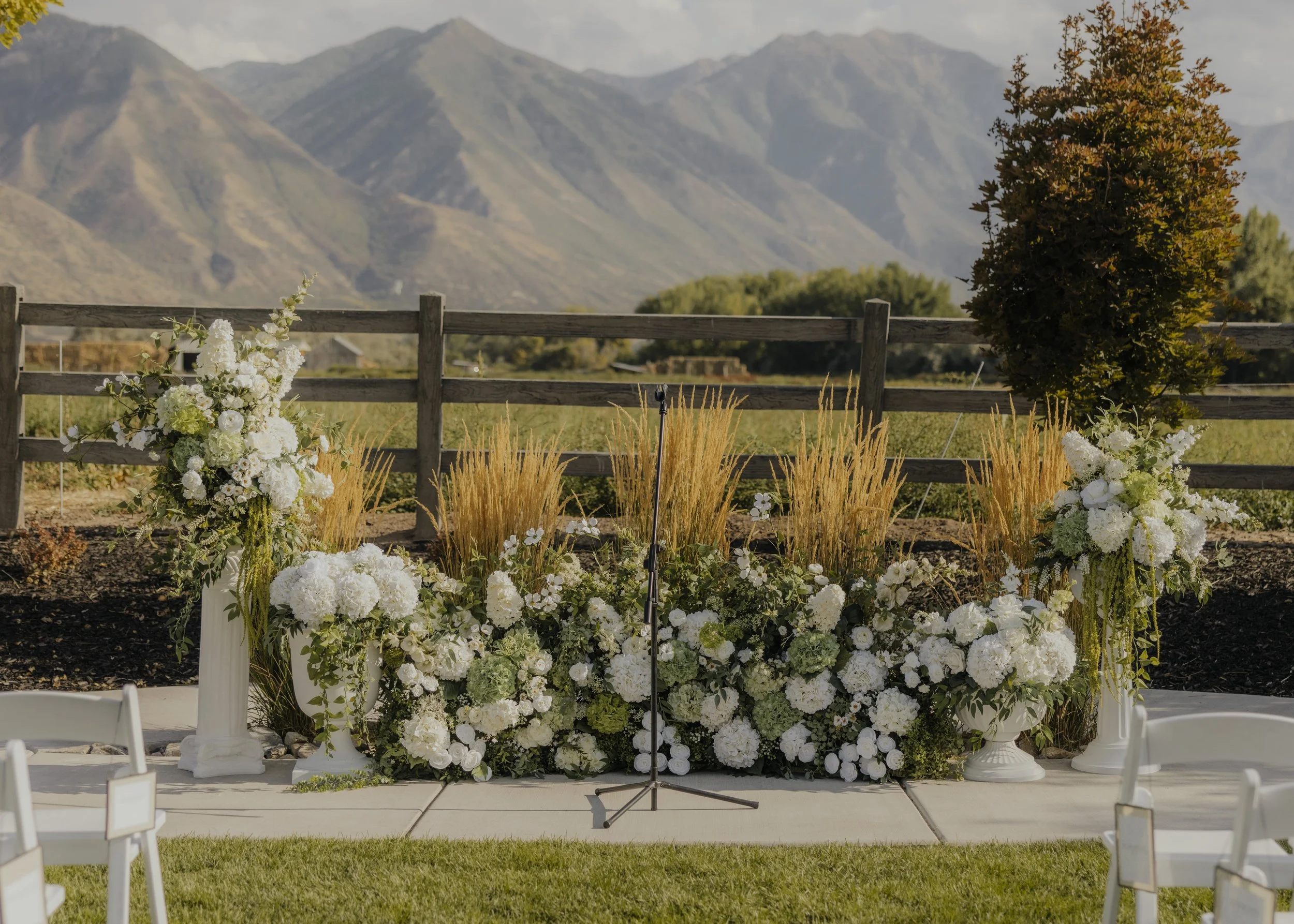 A wedding ceremony setup outdoors with a floral arrangement of white flowers and greenery in front of a mountain range background, with a microphone stand in the center.