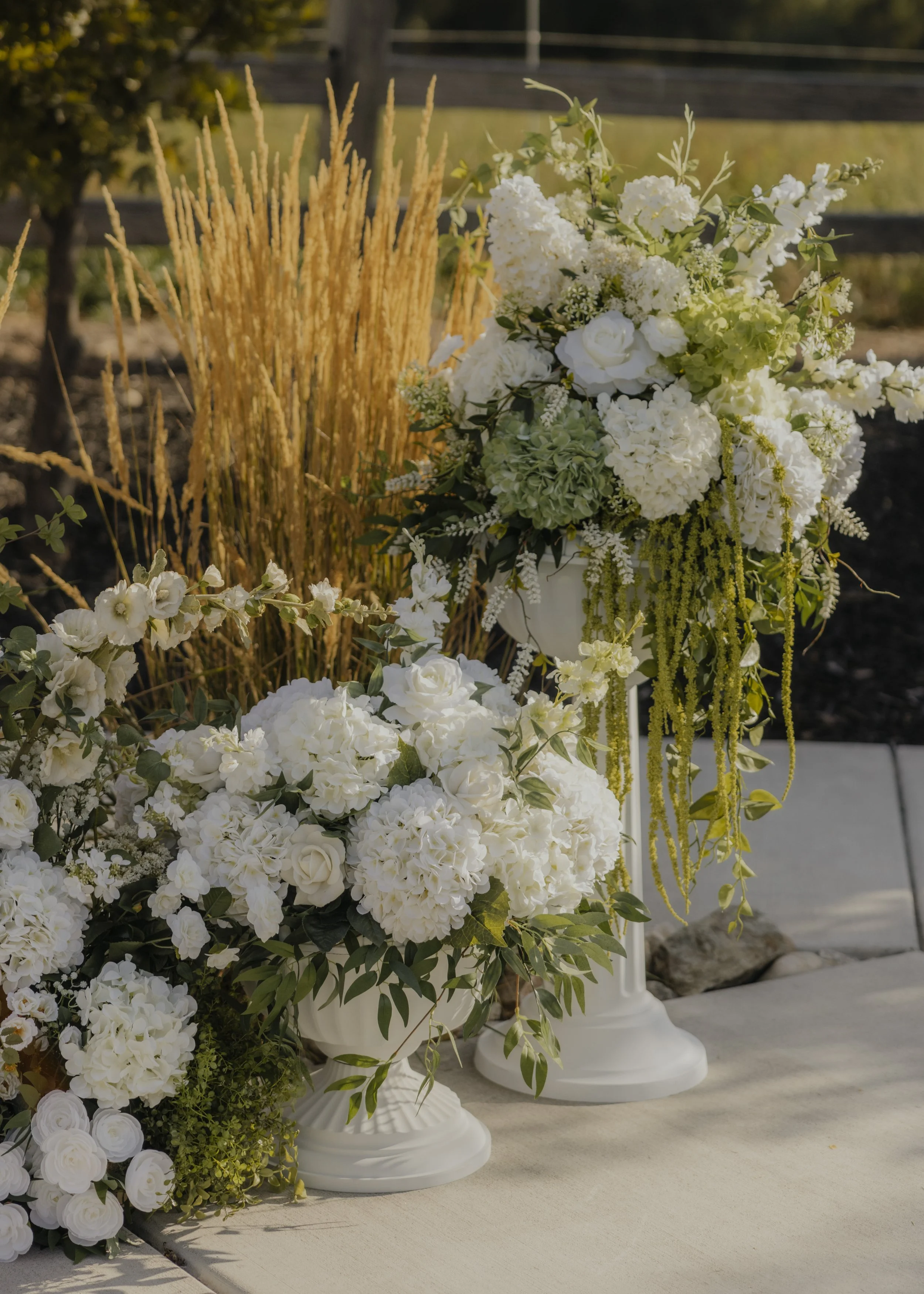 Arrangement of white flowers and greenery in vases on a table outdoors, with wheat stalks and a fence in the background.