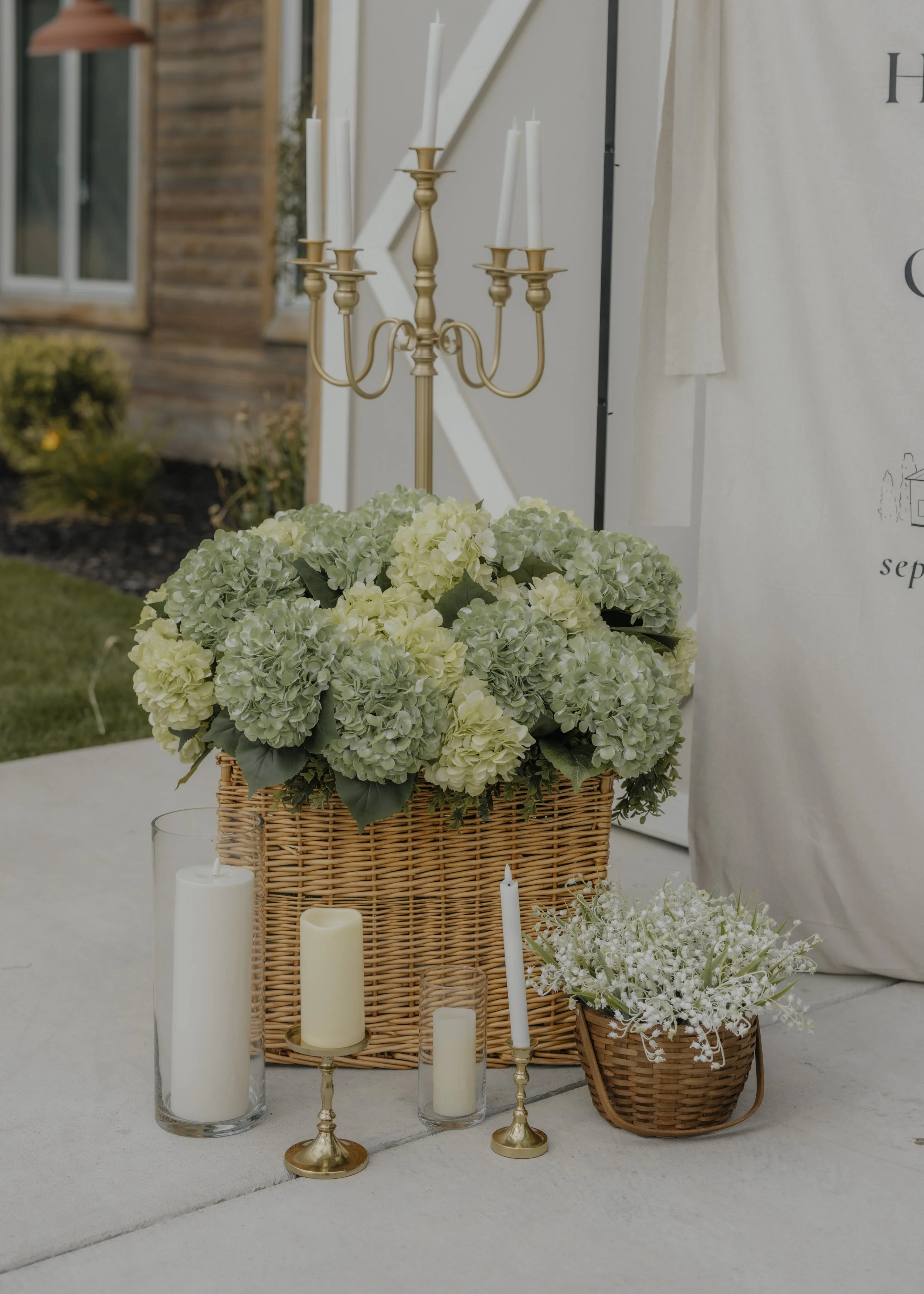 Decorative floral arrangement with white and light green hydrangeas in a wicker basket, surrounded by white candles in glass holders, a gold candelabrum, and a small basket of white flowers, set outdoors with a building and greenery in the background