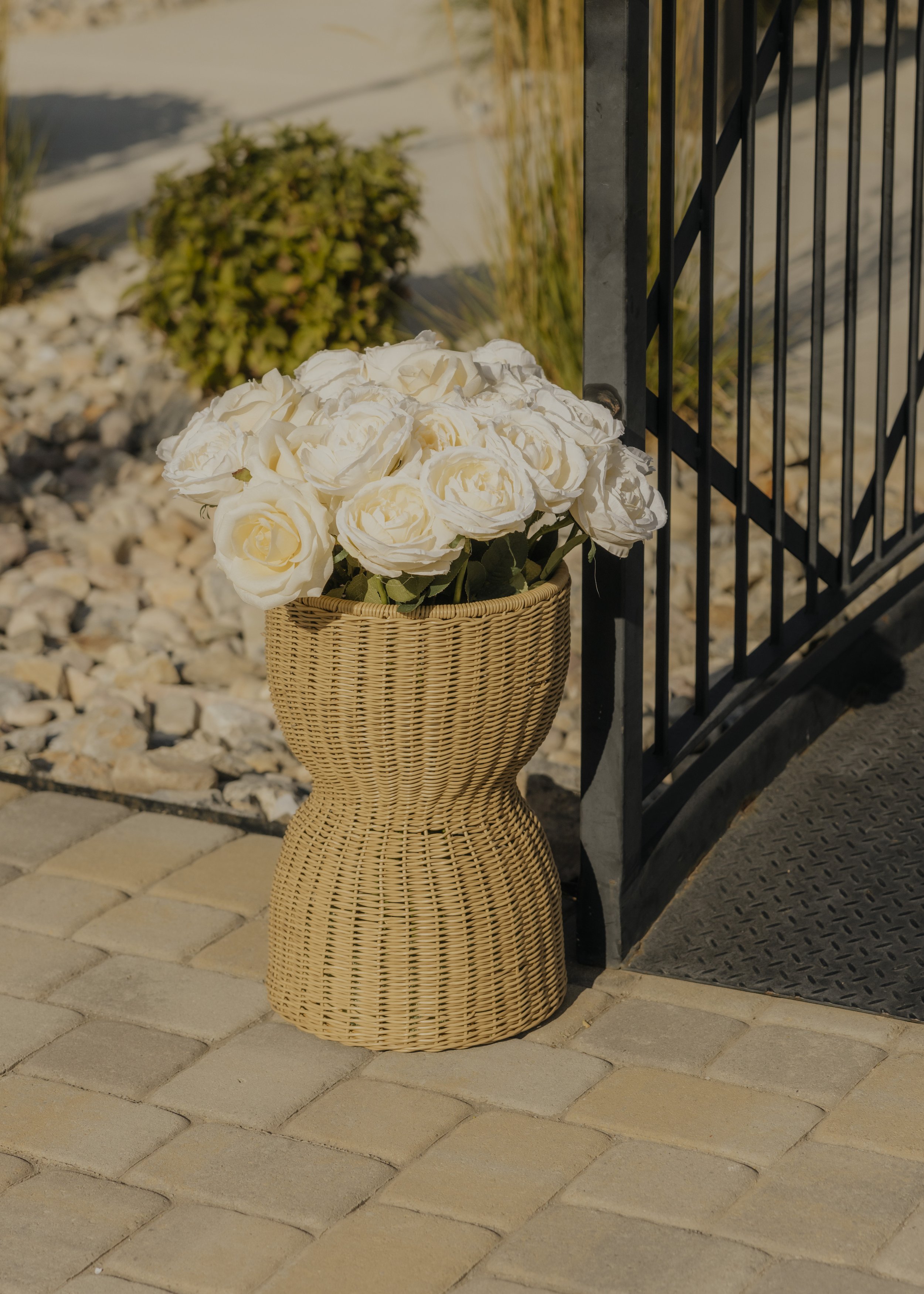 A wicker vase with white roses placed on a paved surface outside near a black metal gate and some decorative rocks and plants.