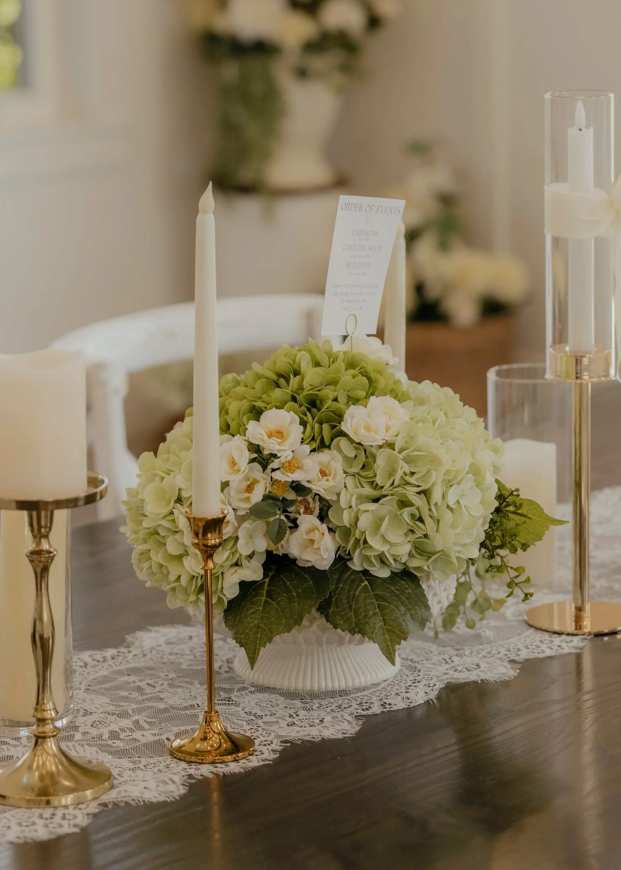 Elegant floral centerpiece with white and green hydrangeas and cream roses on a lace table runner, surrounded by tall gold candlesticks with white candles, set for a formal event or wedding.
