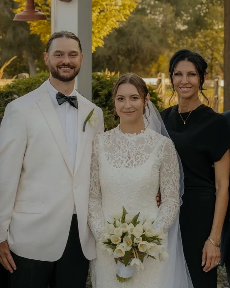 A wedding couple standing outdoors with a woman, all smiling. The groom is wearing a white tuxedo with a black bow tie and a boutonniere. The bride is in a lace wedding gown holding a bouquet of white flowers. The woman is dressed in a black dress with a necklace.