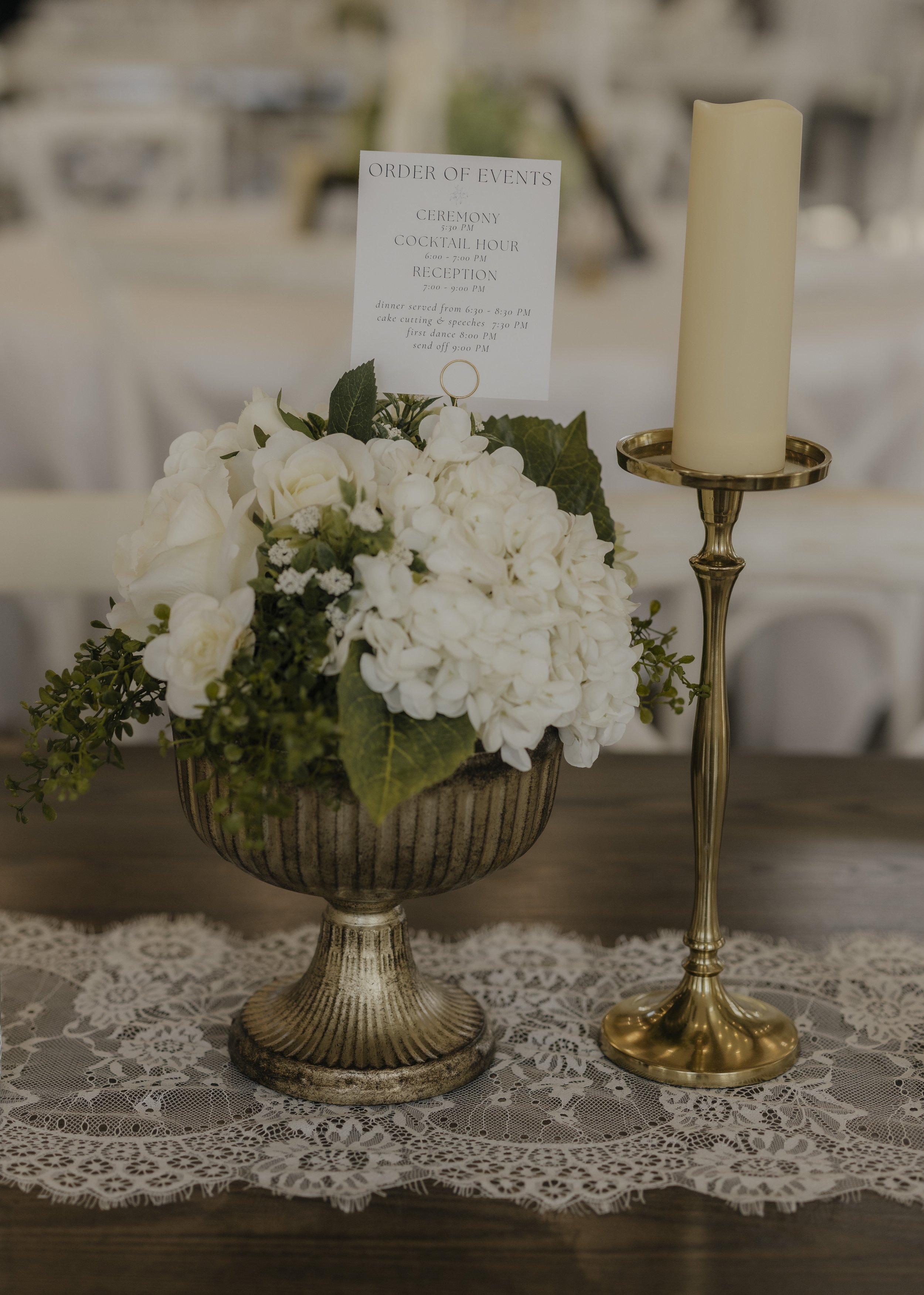 A floral centerpiece with white flowers and green leaves, and a tall white candle on a gold stand, on a lace tablecloth at an event.