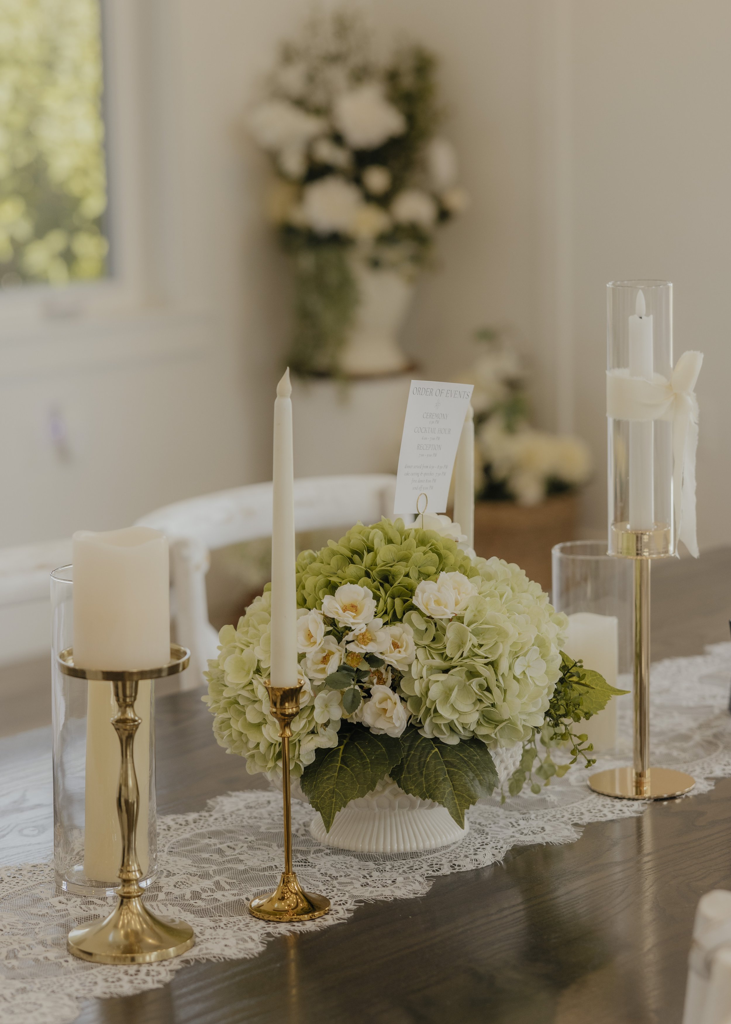 Elegant table centerpiece with white and green flowers, surrounded by white and gold candles, on a lace table runner in a warmly lit room.