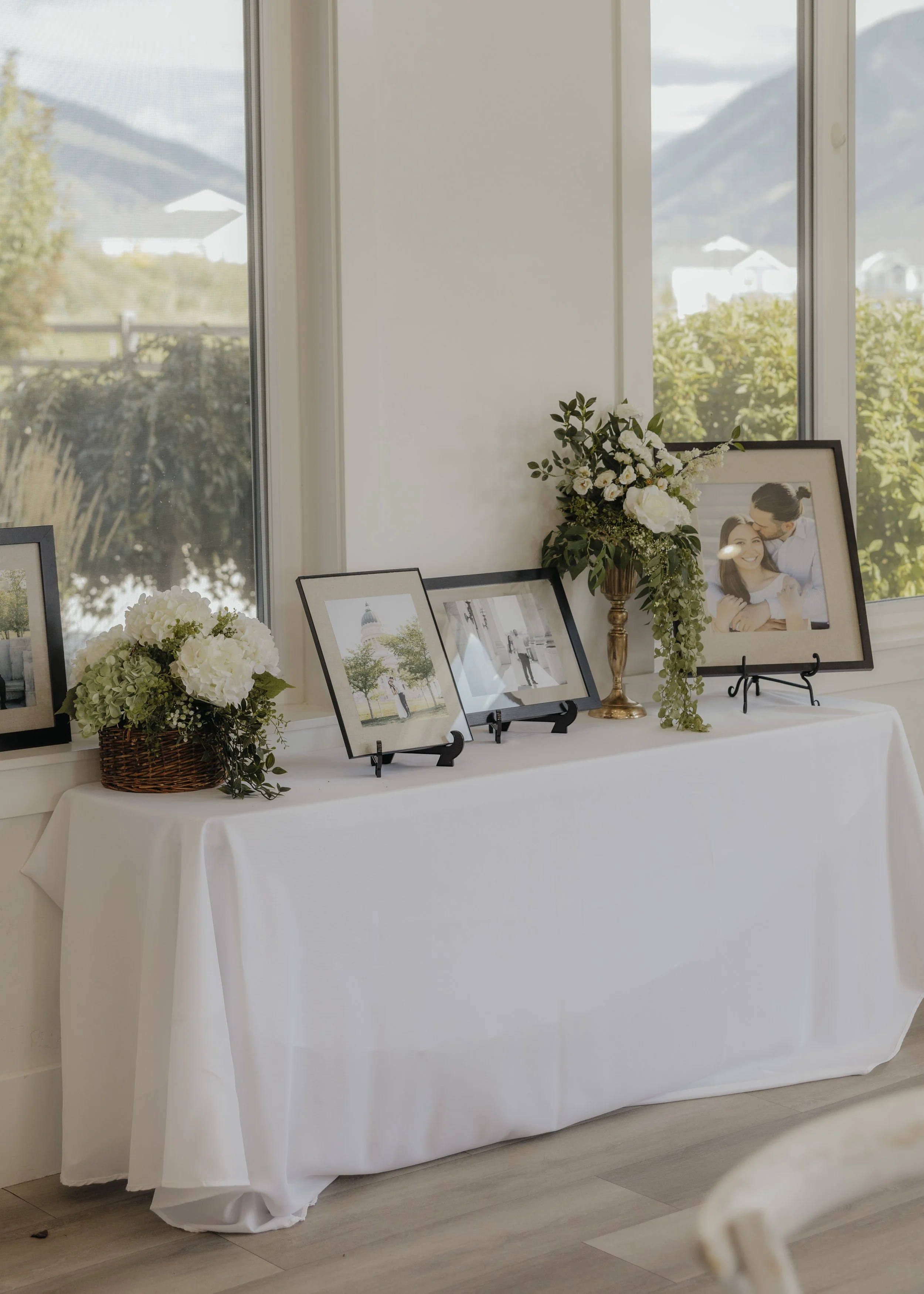 A white tablecloth-covered table with framed photographs and floral arrangements, positioned in front of large windows overlooking a scenic outdoors.