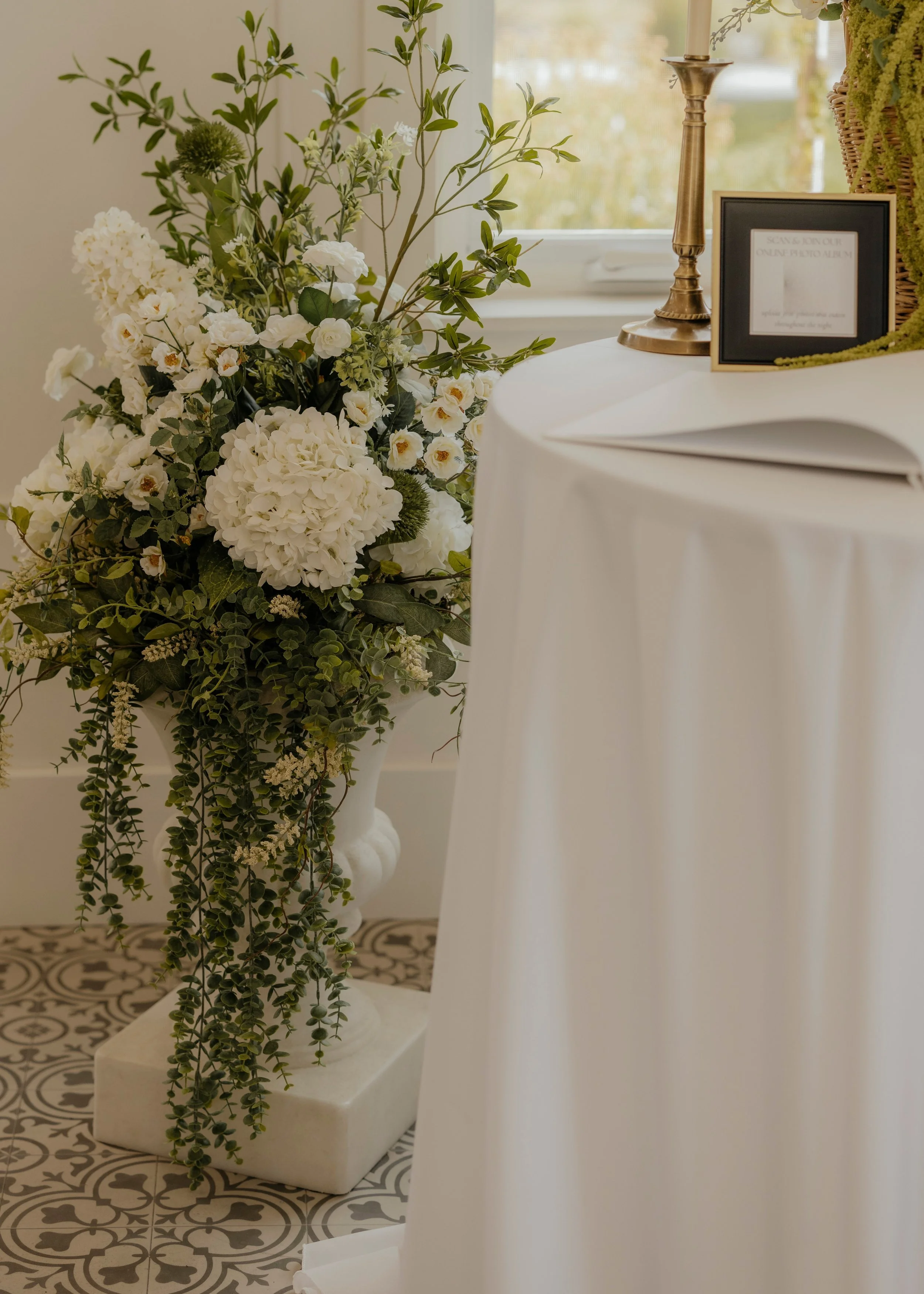 A large floral arrangement with white flowers and green foliage in a tall white urn, placed near a round table with a white tablecloth, a tall brass candlestick, and a small framed sign, in a room with a window and patterned floor tiles.