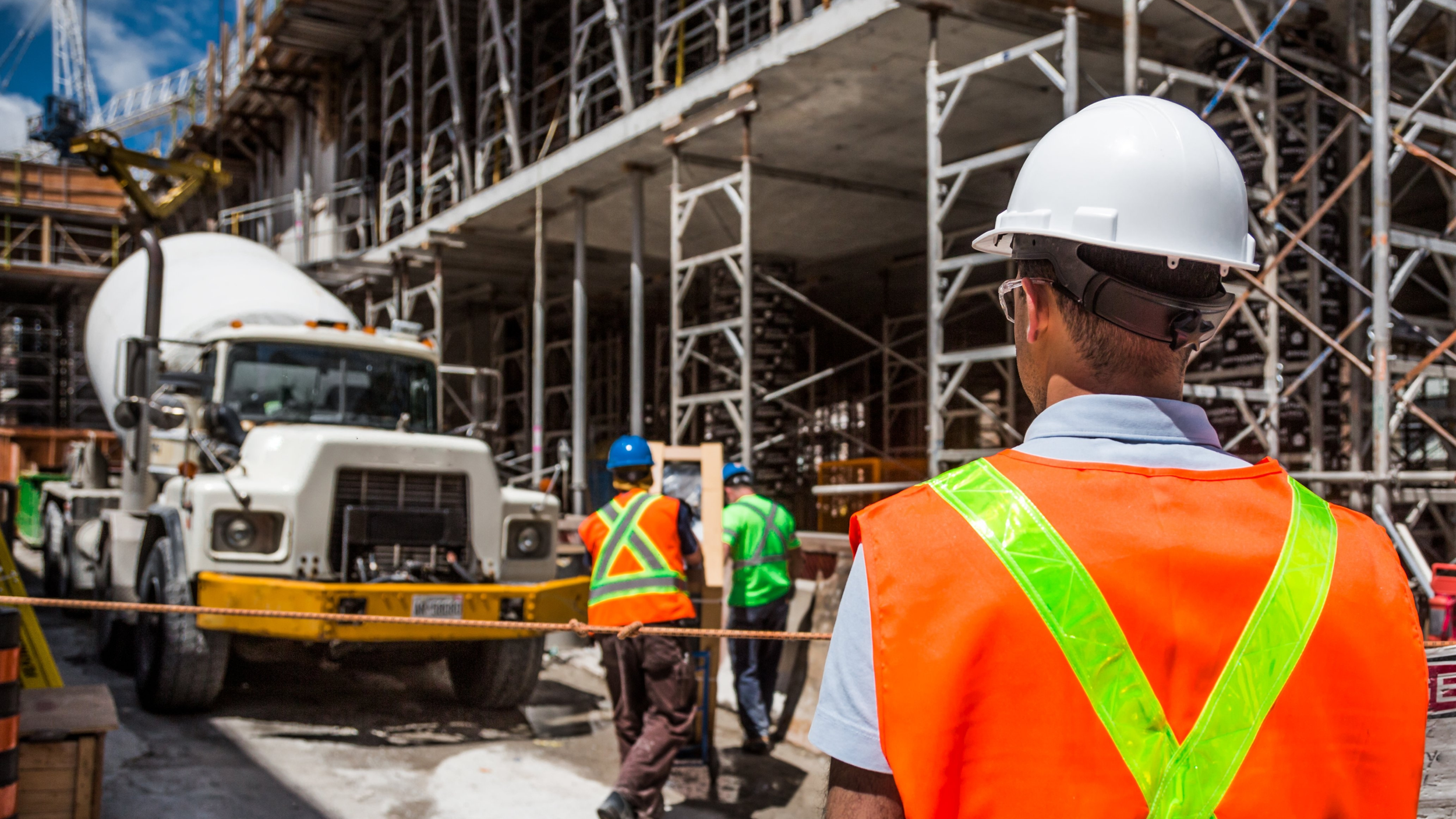 Travailleurs en sécurité portant des gilets orange et des casques de sécurité sur un site de construction, avec un camion-béton et une structure métallique en arrière-plan.