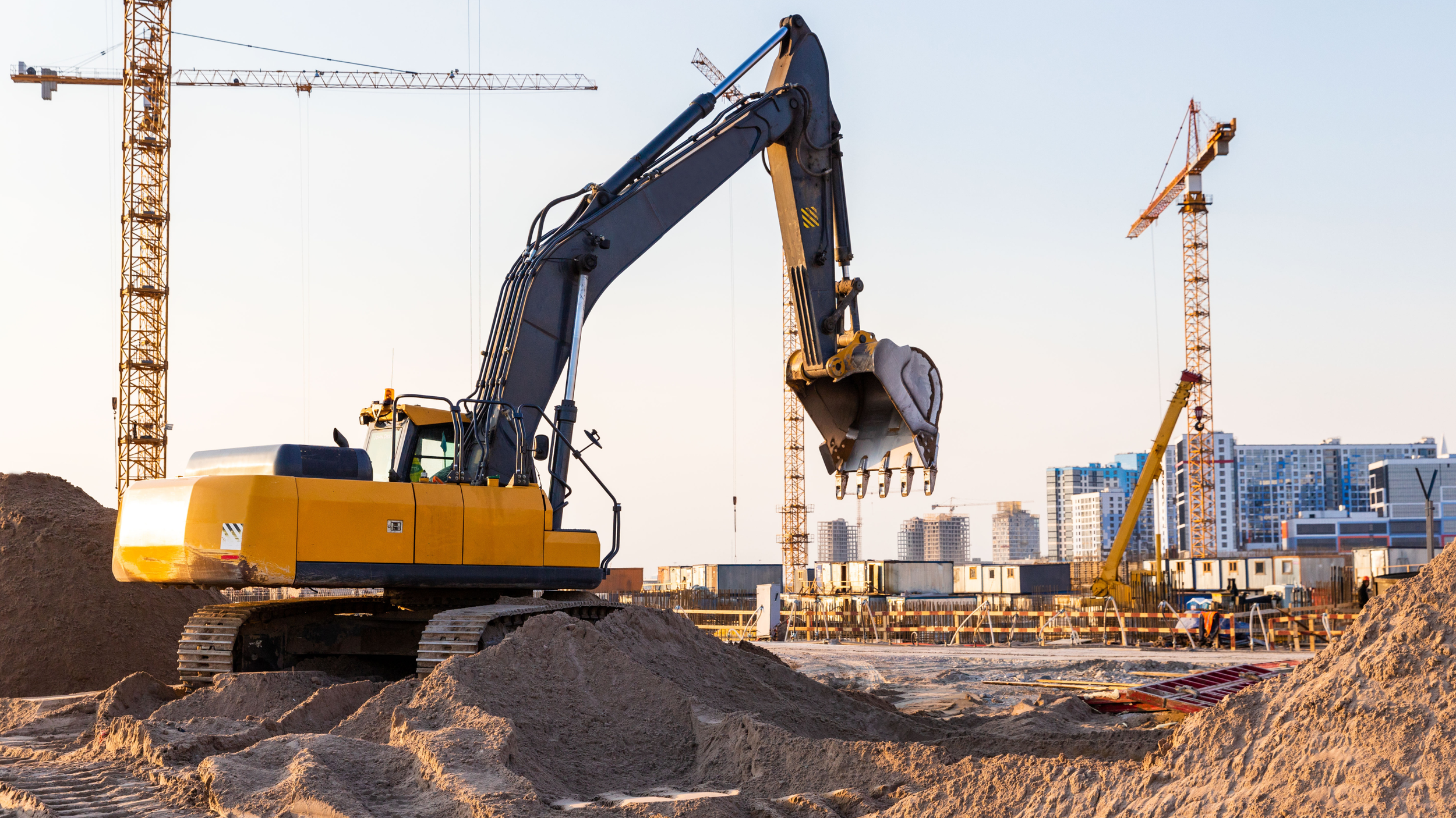 Une pelleteuse jaune sur un chantier de construction, entourée de sables et de tours de cranes en arrière-plan, avec un ciel clair.