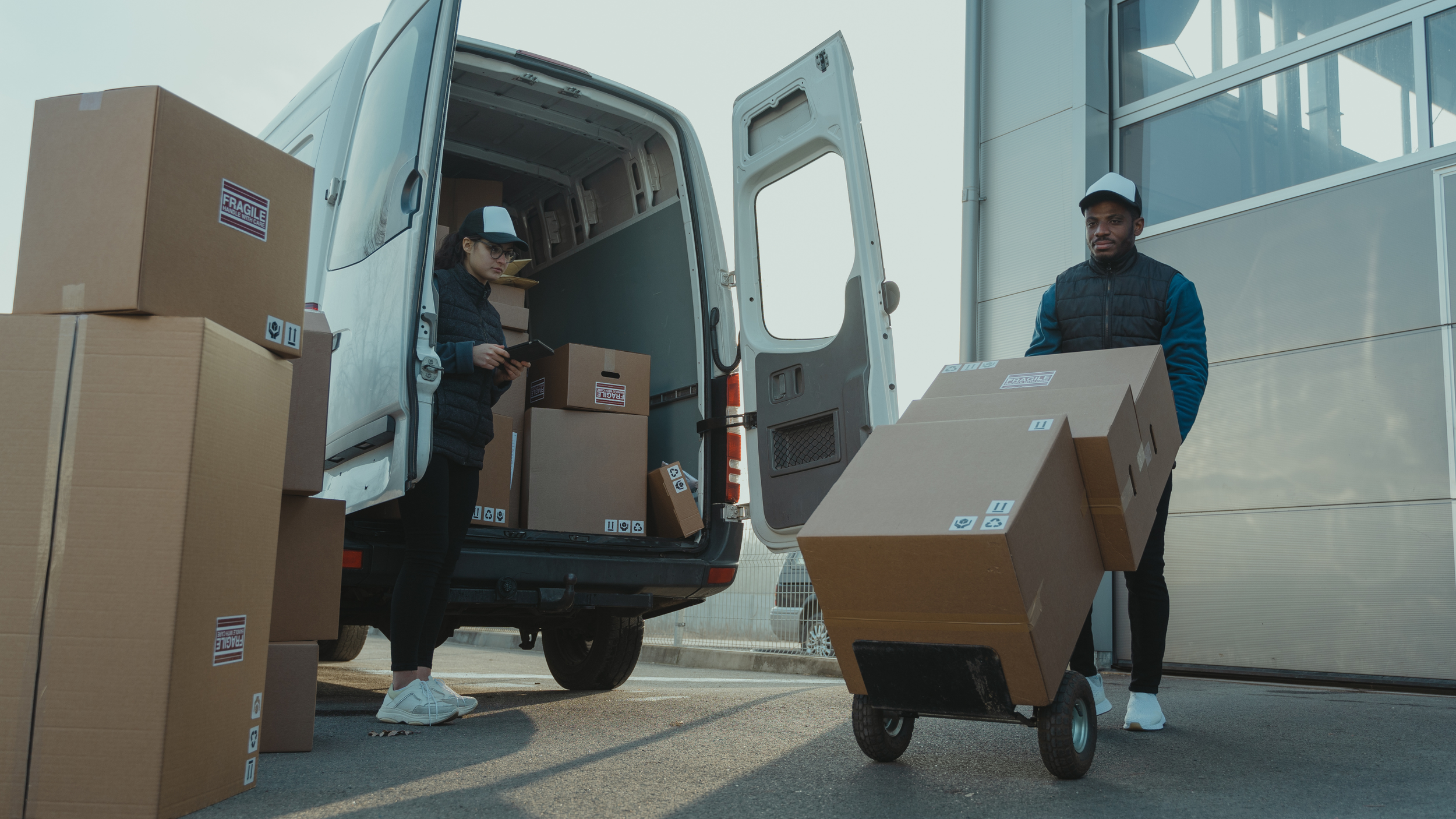 Deux livreurs déchargeant des colis dans une zone extérieure, avec un camion ouvert et un bâtiment moderne en arrière-plan.