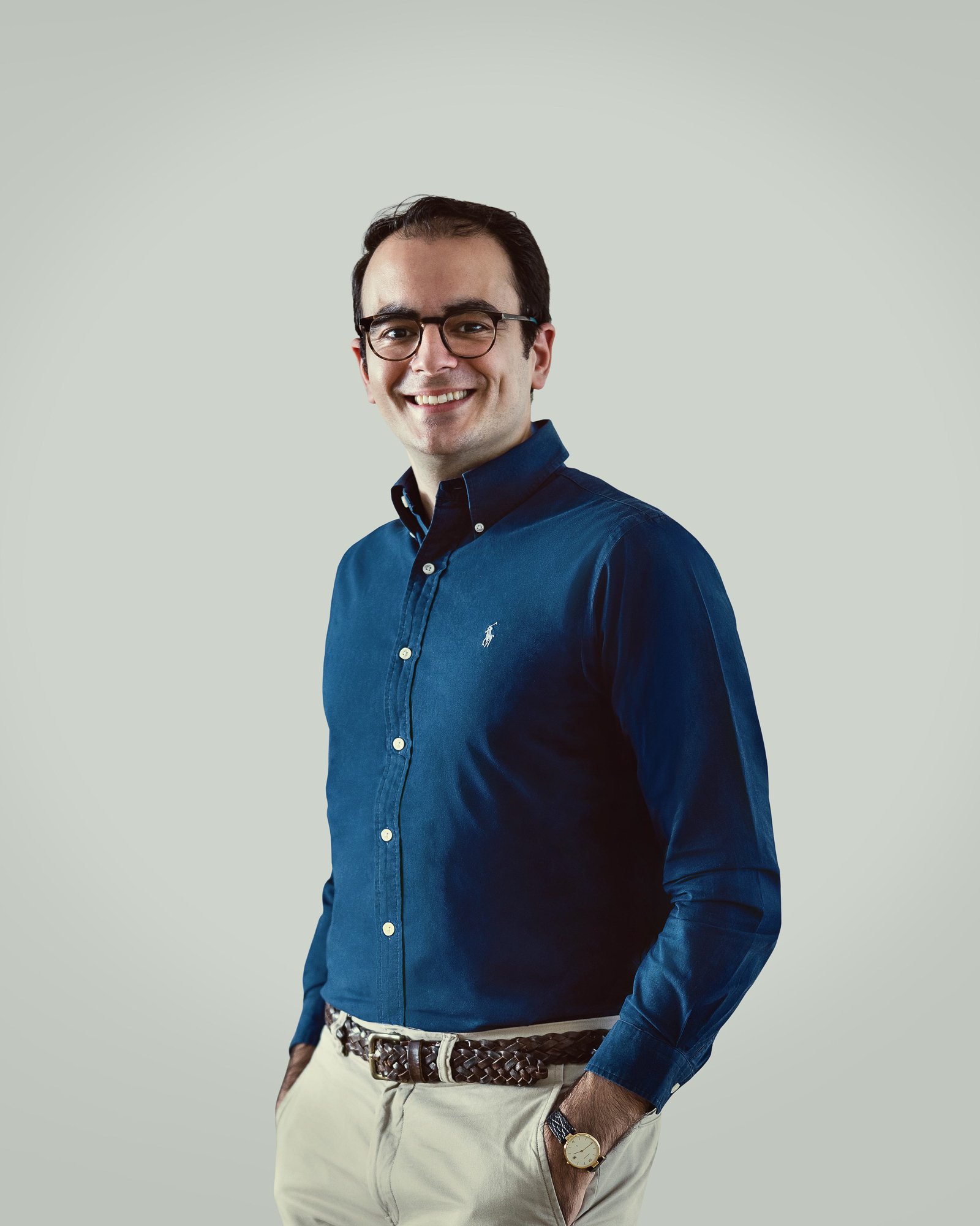 Ein Mann trägt eine dunkelblaue Hemd mit weißen Knöpfen, eine beige Hose, einen braunen geflochtenen Gürtel, eine Armbanduhr und eine Brille, steht vor hellem Hintergrund und lächelt.