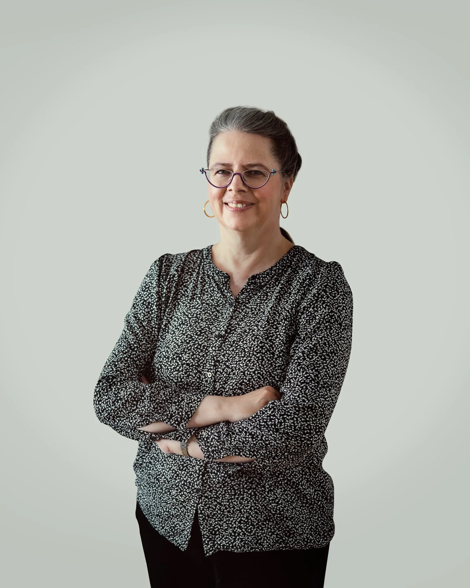 Eine lächelnde Frau mit grauem Haar, Brille und goldenen Ohrringen, die einen schwarzen und weißen gemusterten Satz trägt und die Arme verschränkt hat, vor einem hellgrauen Hintergrund.