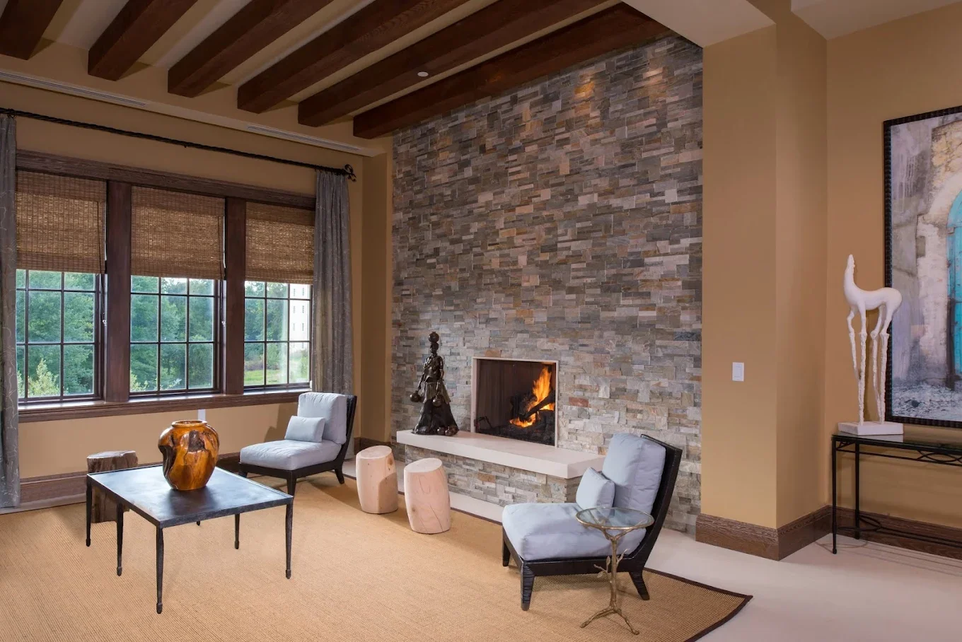 Living room with beige walls, large windows with bamboo shades, a stone fireplace, two white armchairs, a black side table with a ceramic pitcher, a small sculpture, and a white sculpture of a horse on a black table.